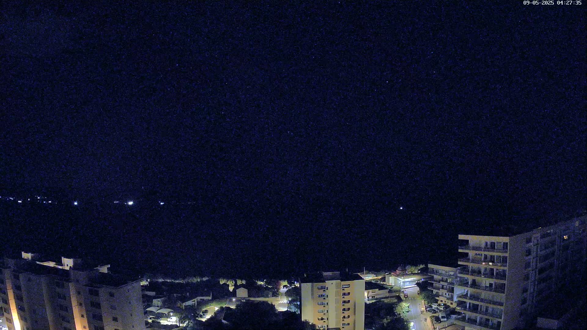 A nighttime view shows a dark, starry sky above a coastal town with several multi-story buildings illuminated and a dark body of water in the distance.
