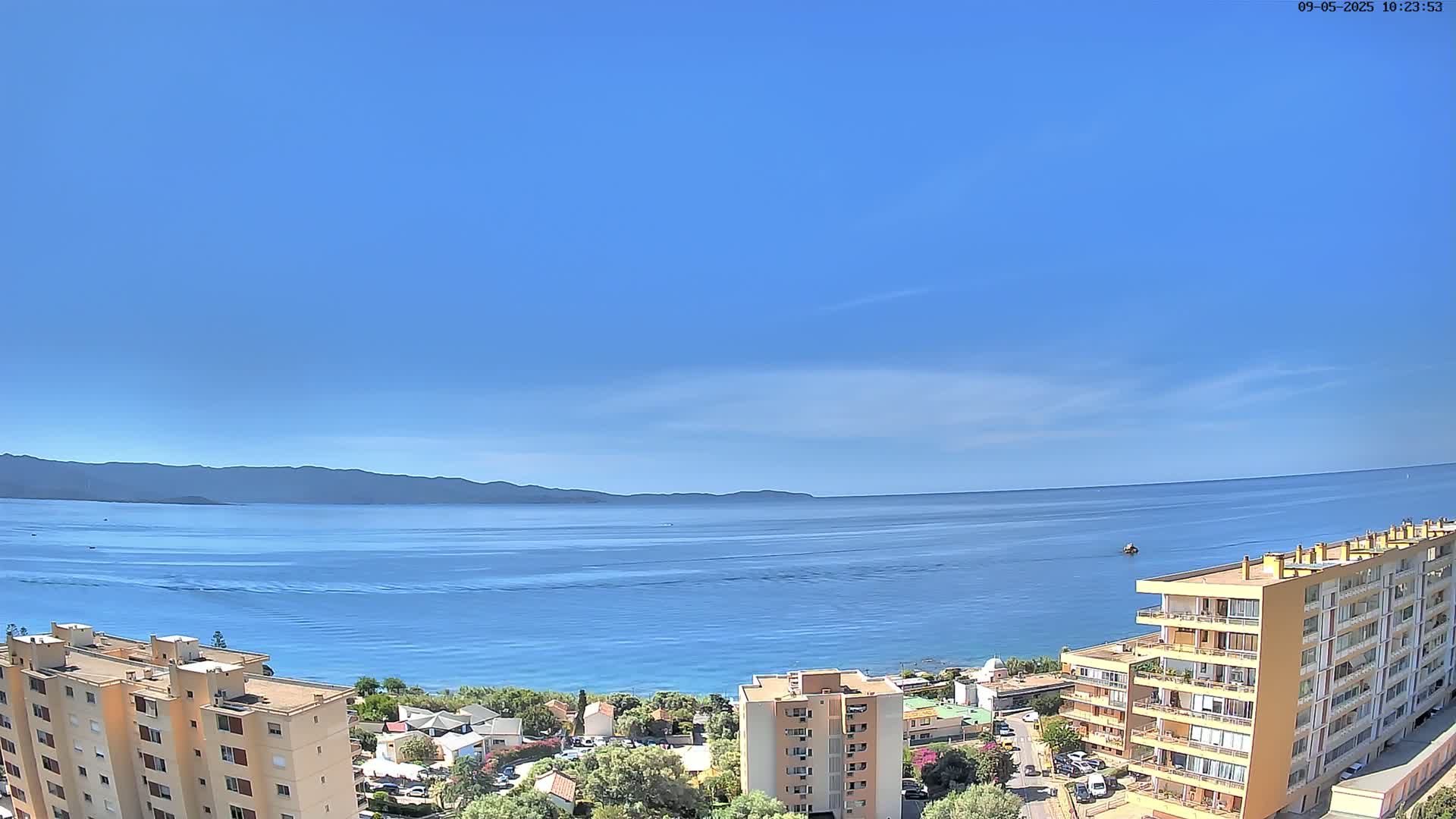 A mostly clear blue sky overlooks a calm blue sea, with distant mountains and several apartment buildings in the foreground.