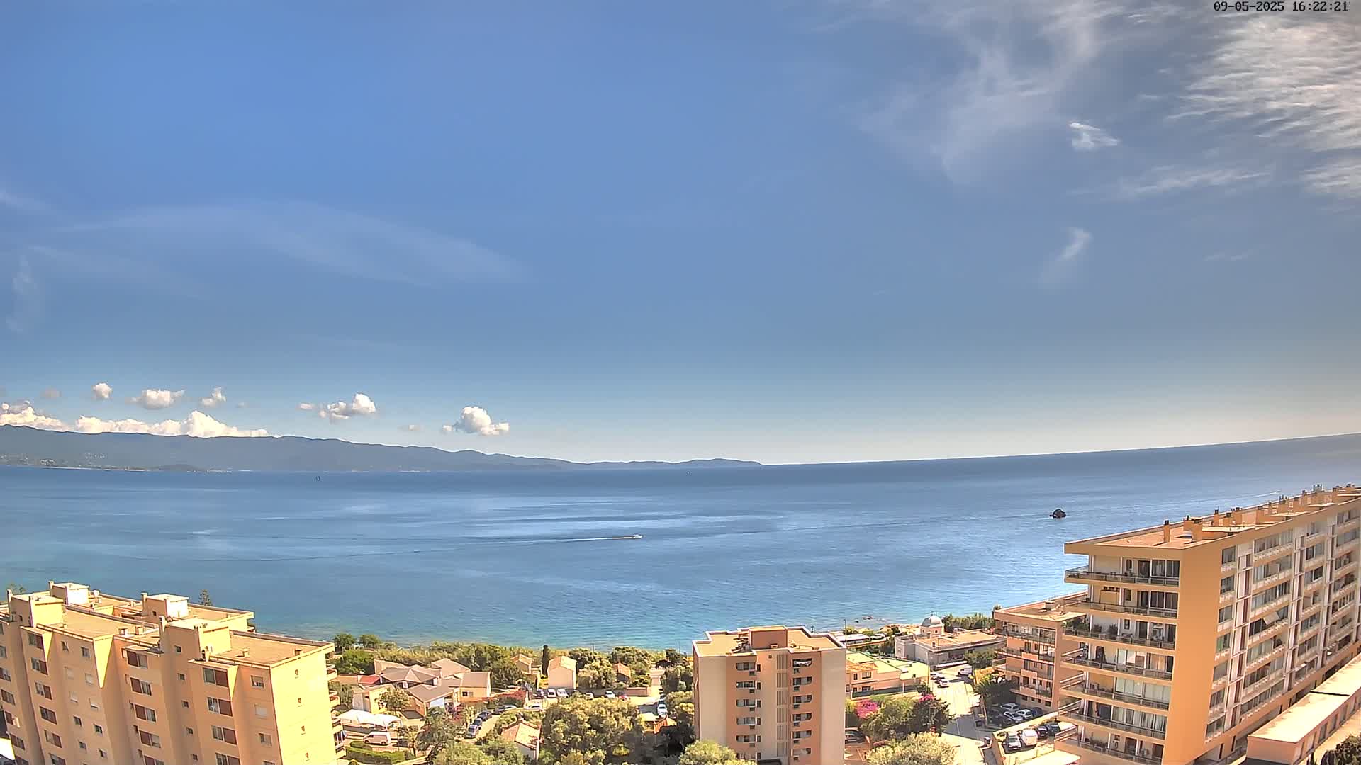 A sunny day shows a calm blue sea, a distant mountain range, and several multi-story buildings on a coastal hillside.