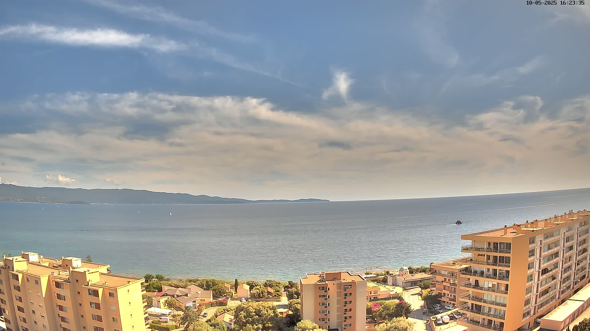 A partly cloudy sky overlooks a calm sea with a distant mountain range, and several multi-story buildings are situated near the shore.