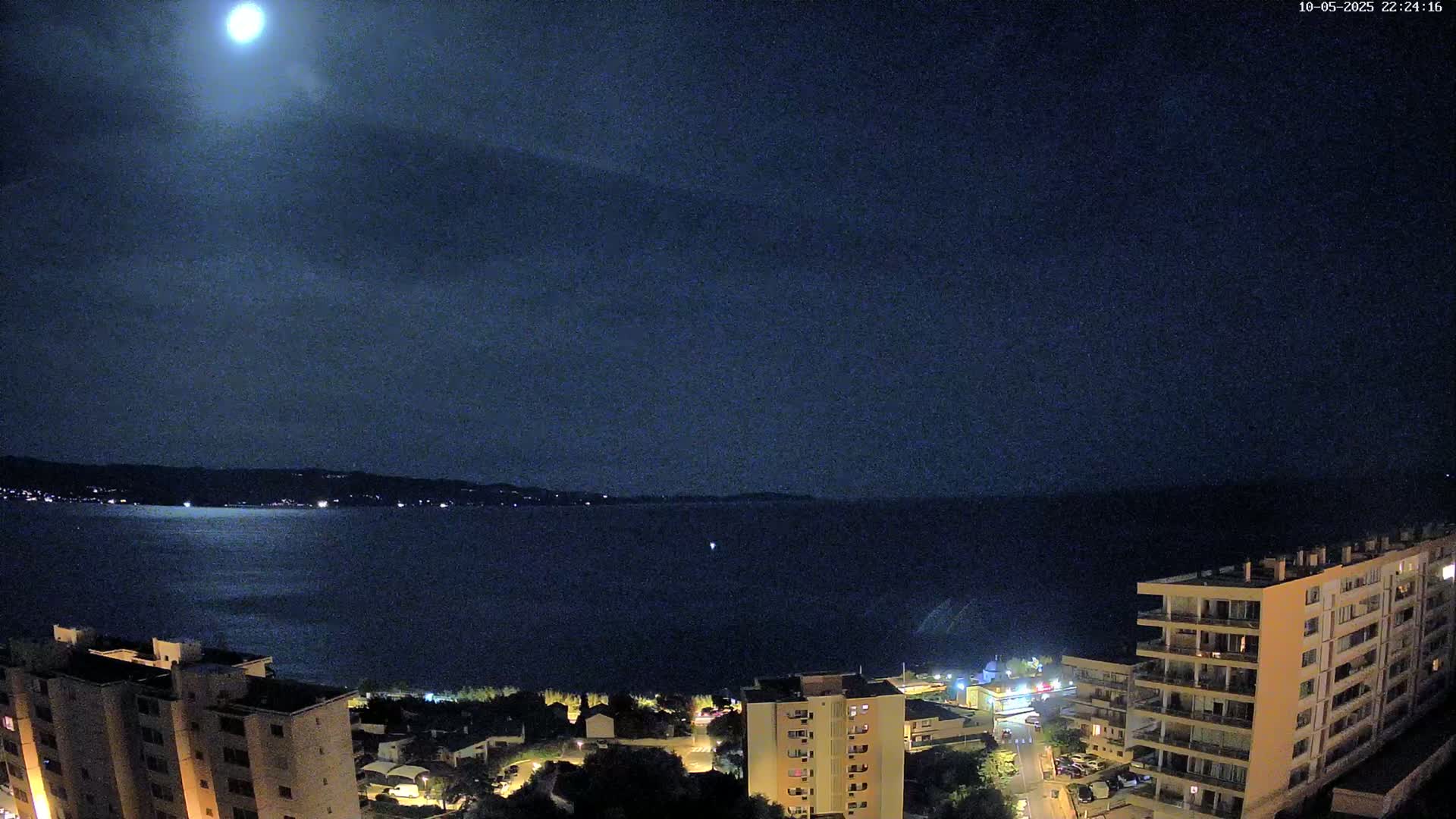 A bright moon illuminates a dark nighttime scene of a calm body of water, bordered by a shoreline with buildings and trees under a mostly clear sky.