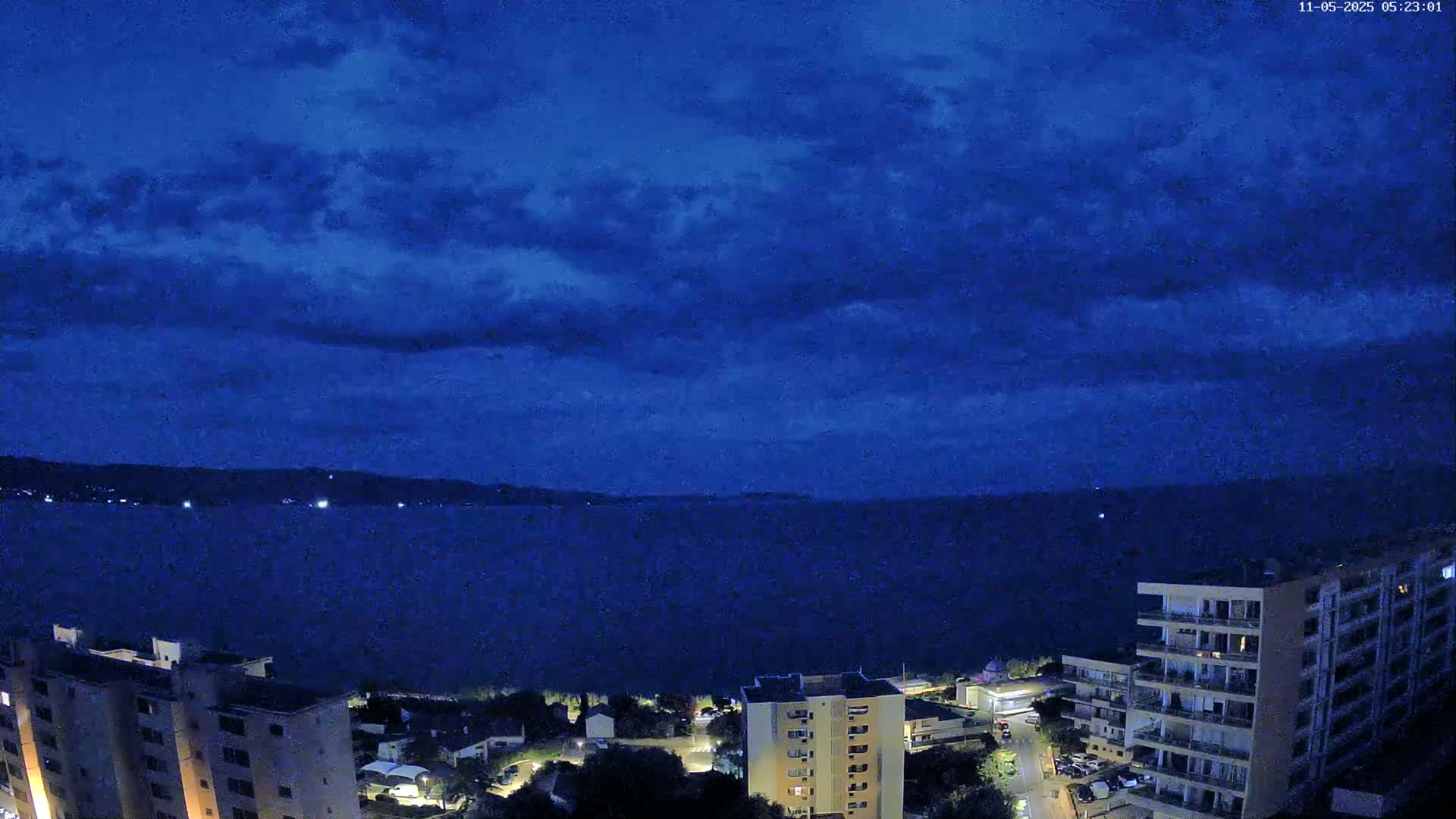 A nighttime view of a dark blue sea under a mostly cloudy sky shows a shoreline with several multi-story buildings.