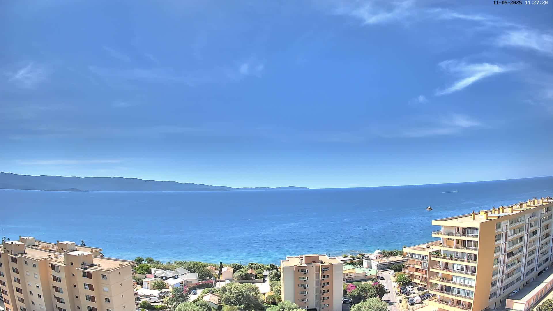 A mostly clear blue sky overlooks a calm blue sea and a coastal town with several multi-story buildings.