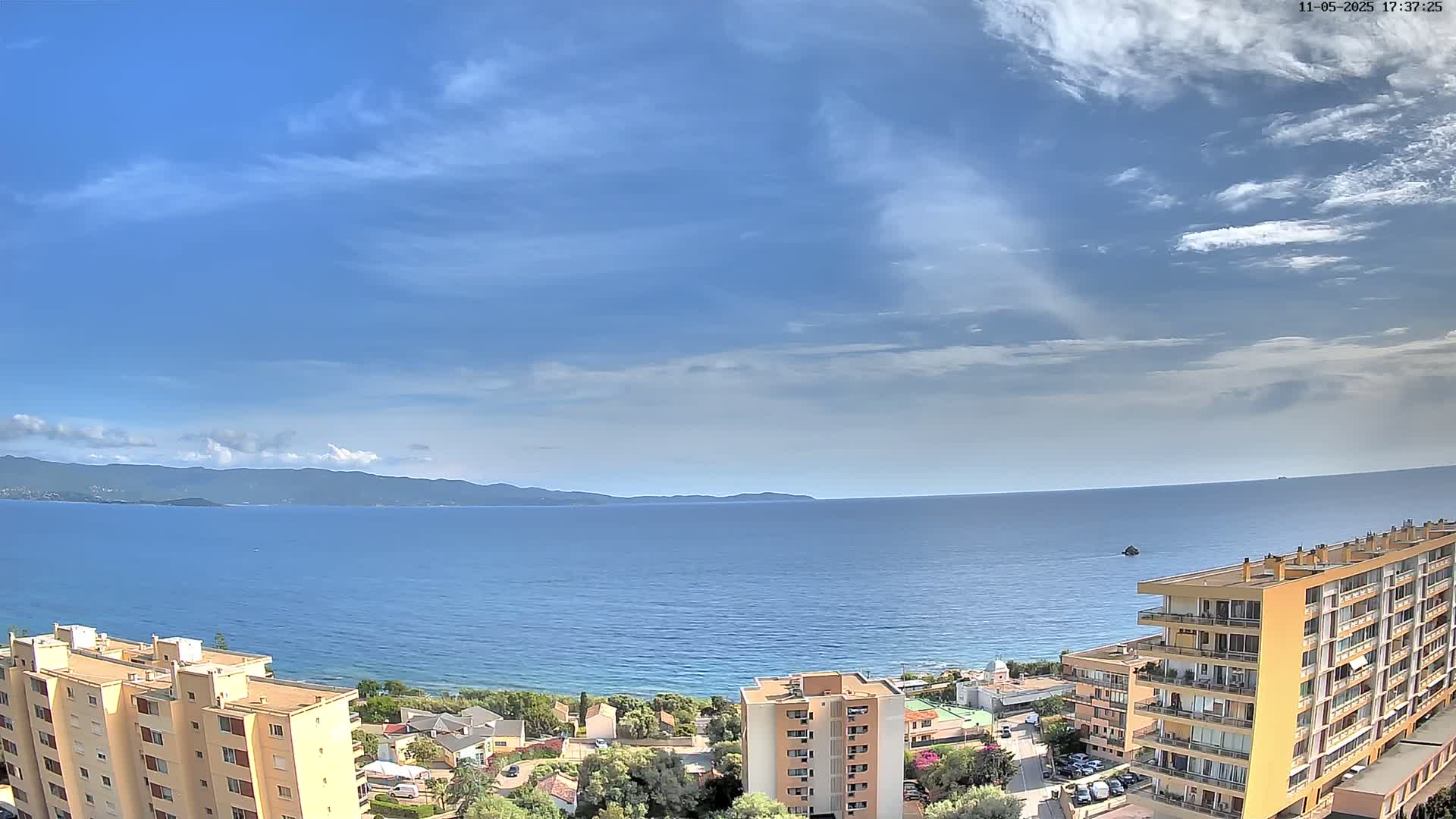 A partly cloudy sky overlooks a calm blue sea, mountains in the distance, and several multi-story buildings.