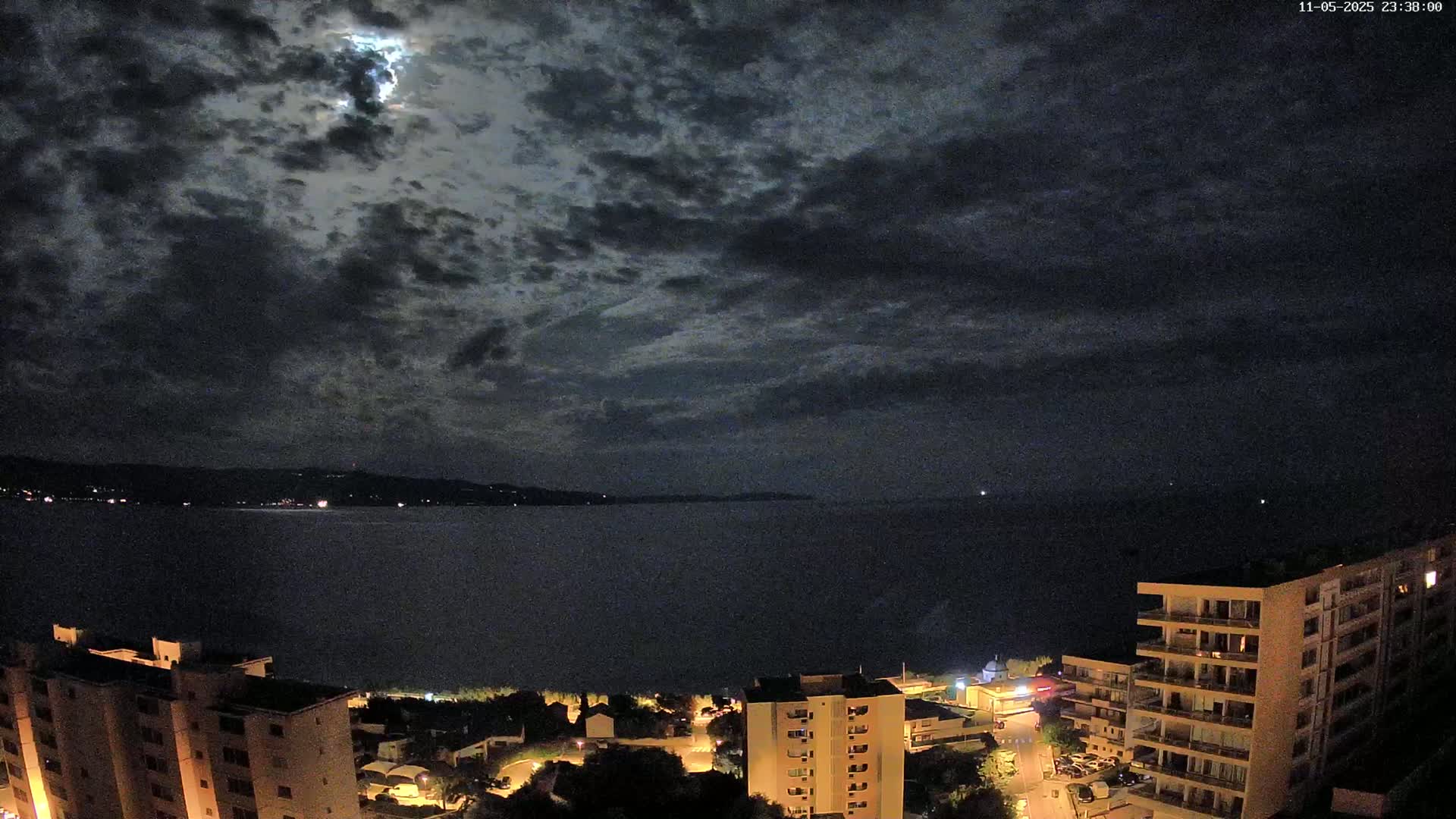 A nighttime coastal scene shows a partly cloudy sky with a bright moon, a dark body of water, and illuminated buildings along the shoreline.