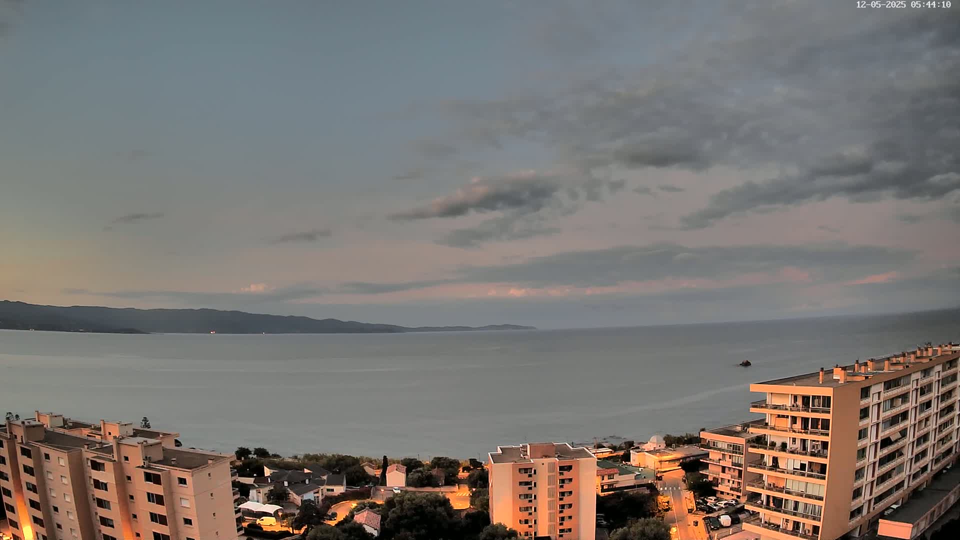 A calm sea under a cloudy, dusky sky is viewed from above a coastal town with several multi-story buildings illuminated at night.