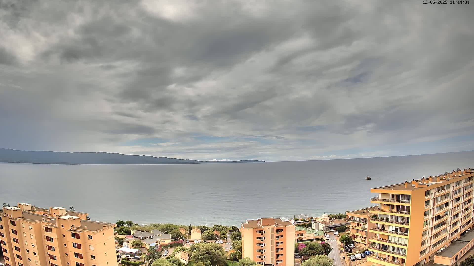 A calm sea stretches to a distant mountain range under a mostly cloudy sky, with apartment buildings visible in the foreground.