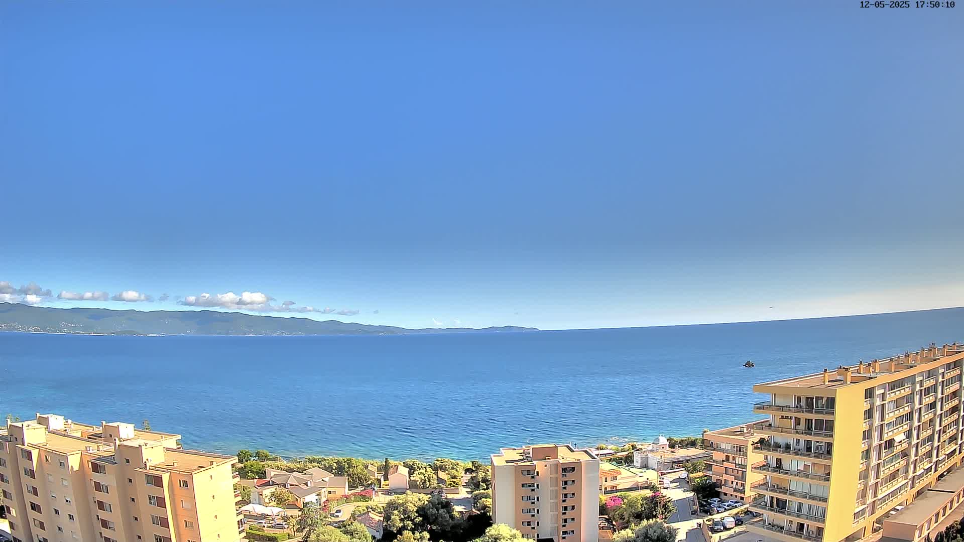 A clear blue sky overlooks a calm blue sea, with a distant mountain range and coastal buildings in the foreground.