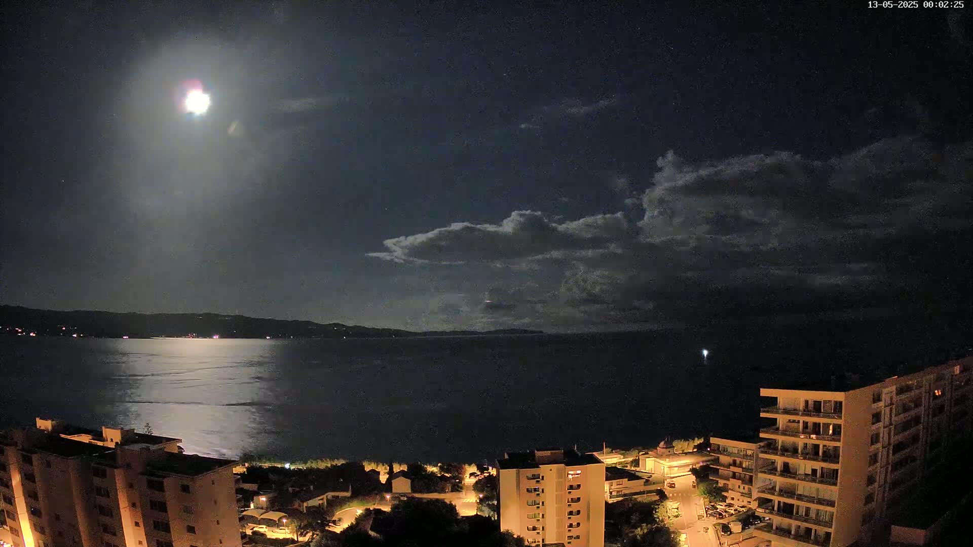 A bright moon shines in a partly cloudy night sky over a calm body of water, with illuminated buildings and streets visible along the shoreline.