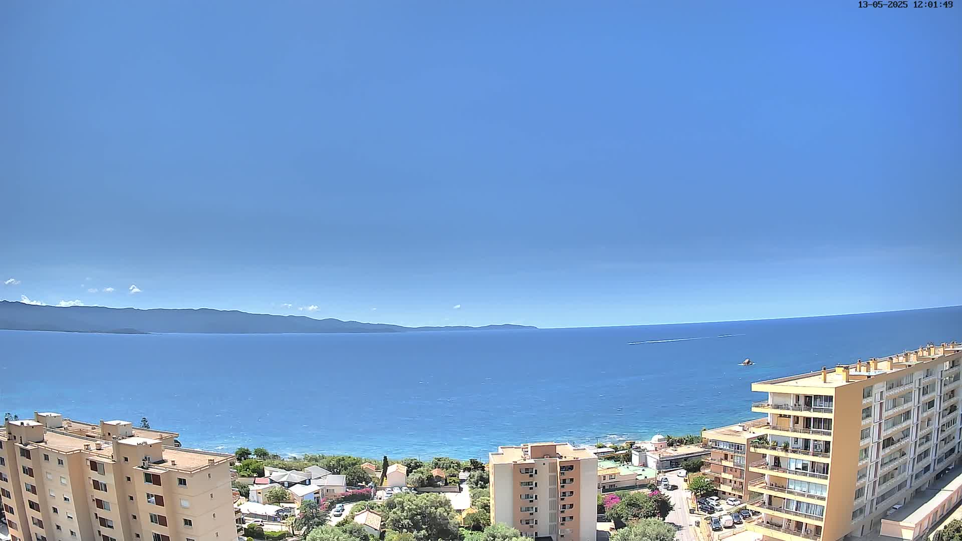 A clear blue sky overlooks a calm blue sea, with a distant mountain range and several buildings along the shoreline.