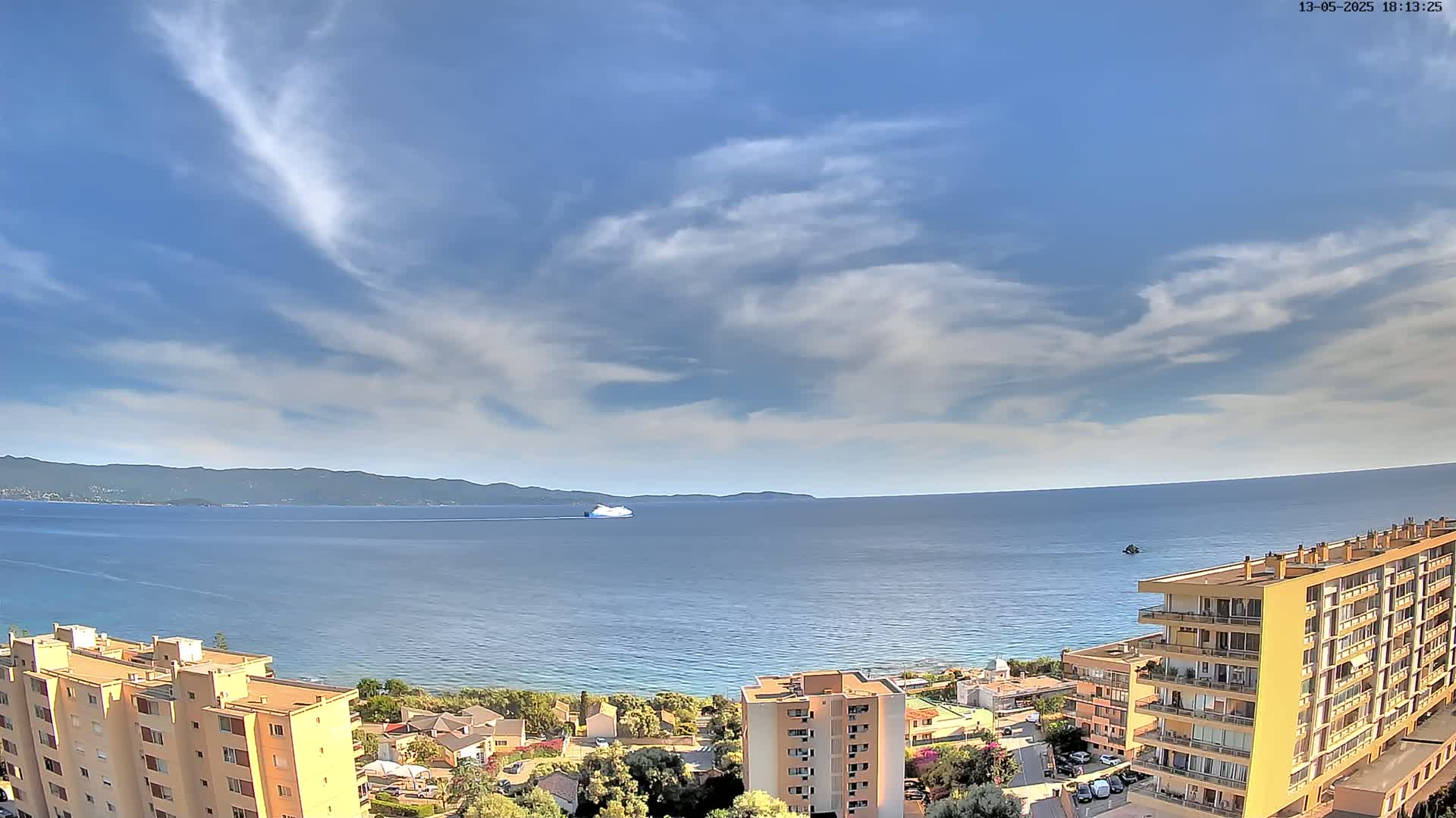 A ferry is visible on a calm sea under a partly cloudy sky, with apartment buildings and coastal vegetation in the foreground.