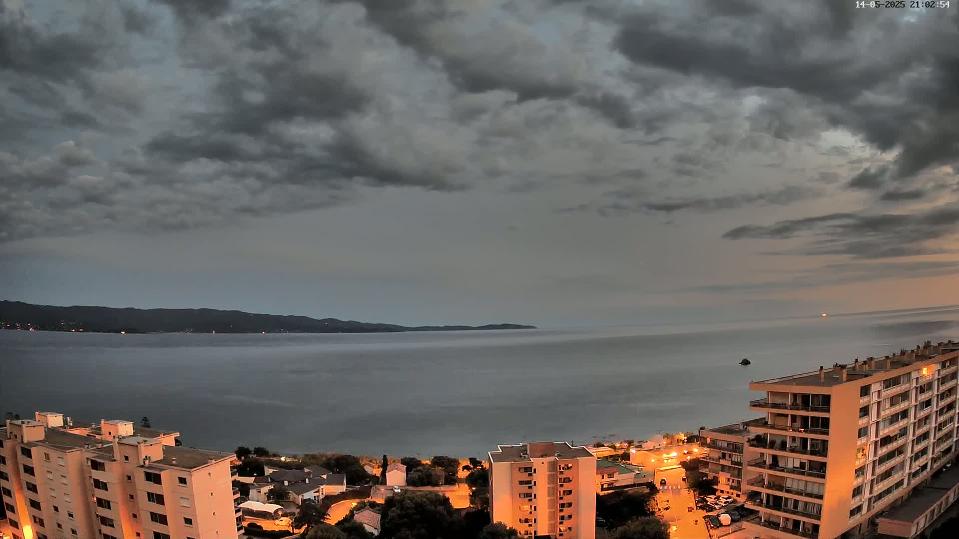 A coastal town at dusk under a mostly cloudy sky overlooks a calm sea and distant mountains.
