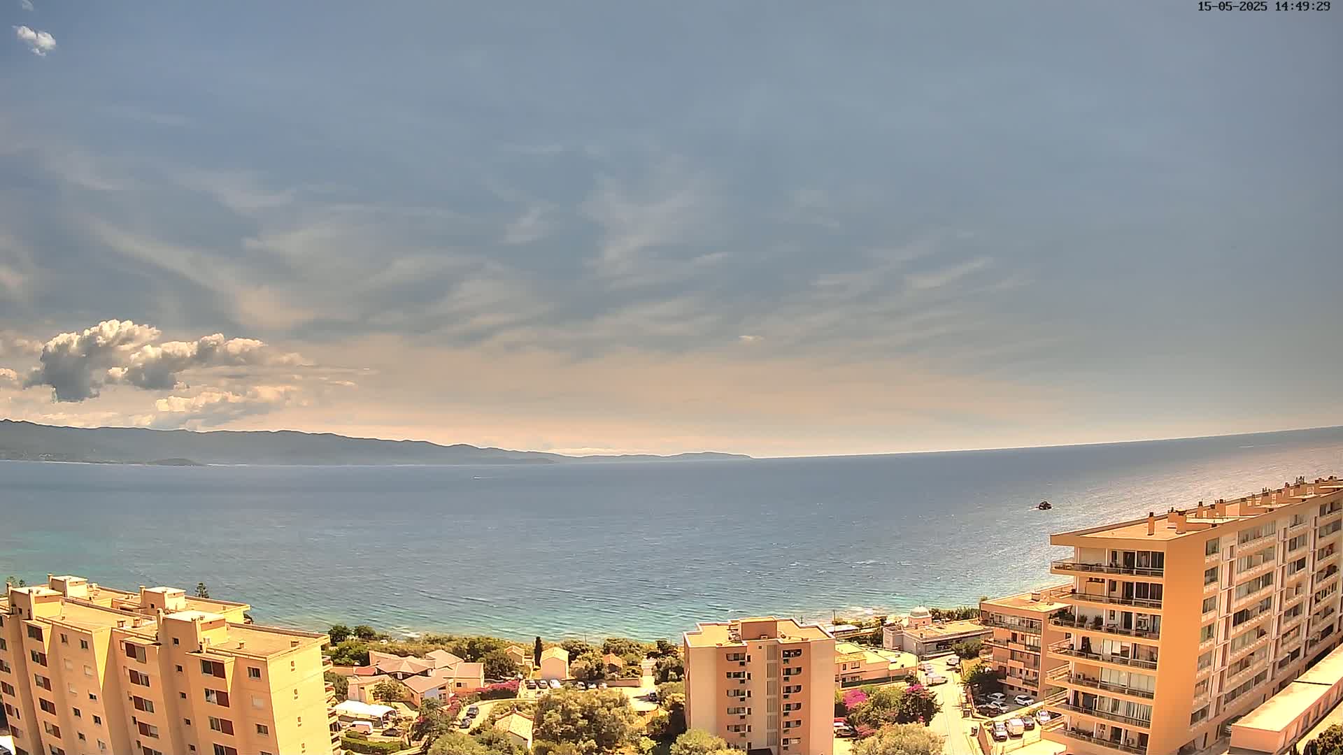 A partly cloudy sky overlooks a calm sea, with a distant mountain range and several multi-story buildings situated on the shoreline.