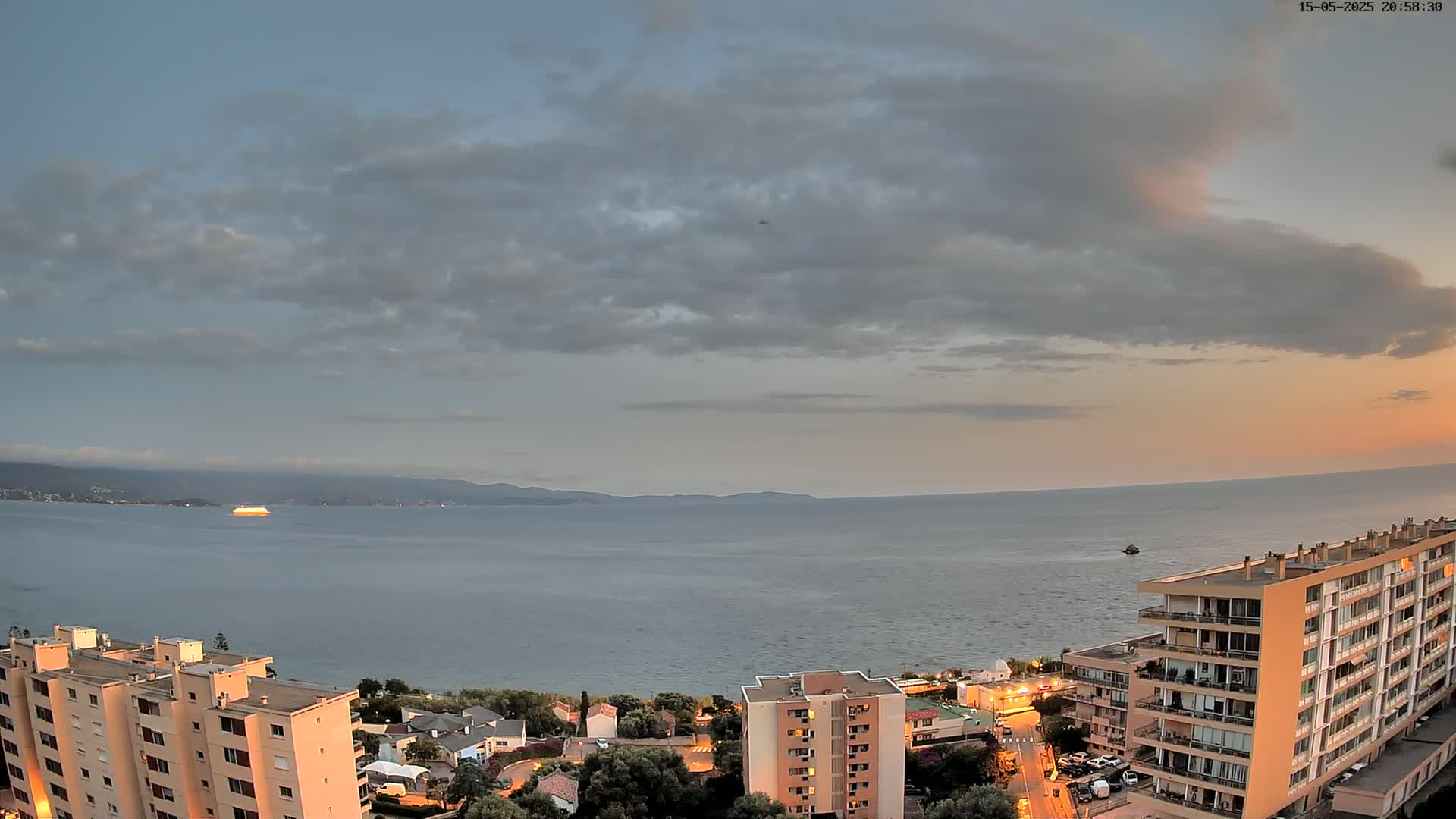A coastal city scene at sunset under a partly cloudy sky shows a body of water with a large vessel, and several apartment buildings with lights on.