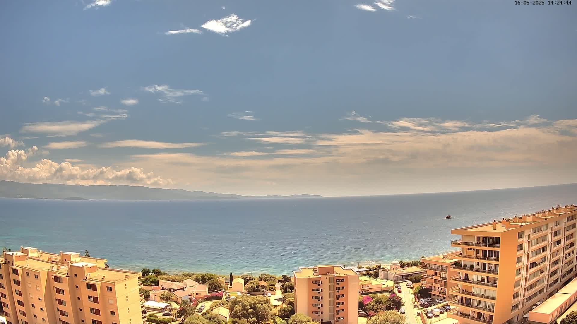 A mostly sunny day reveals a coastal scene with calm blue water, distant mountains, and several multi-story buildings nestled along the shoreline.