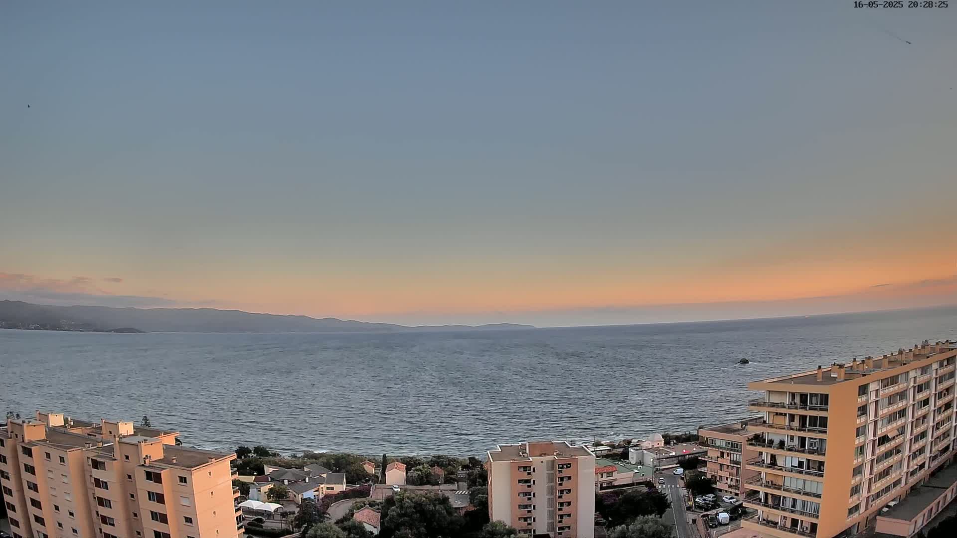 A calm sea with a pastel sunset over distant mountains is viewed from above a coastal town with several apartment buildings.
