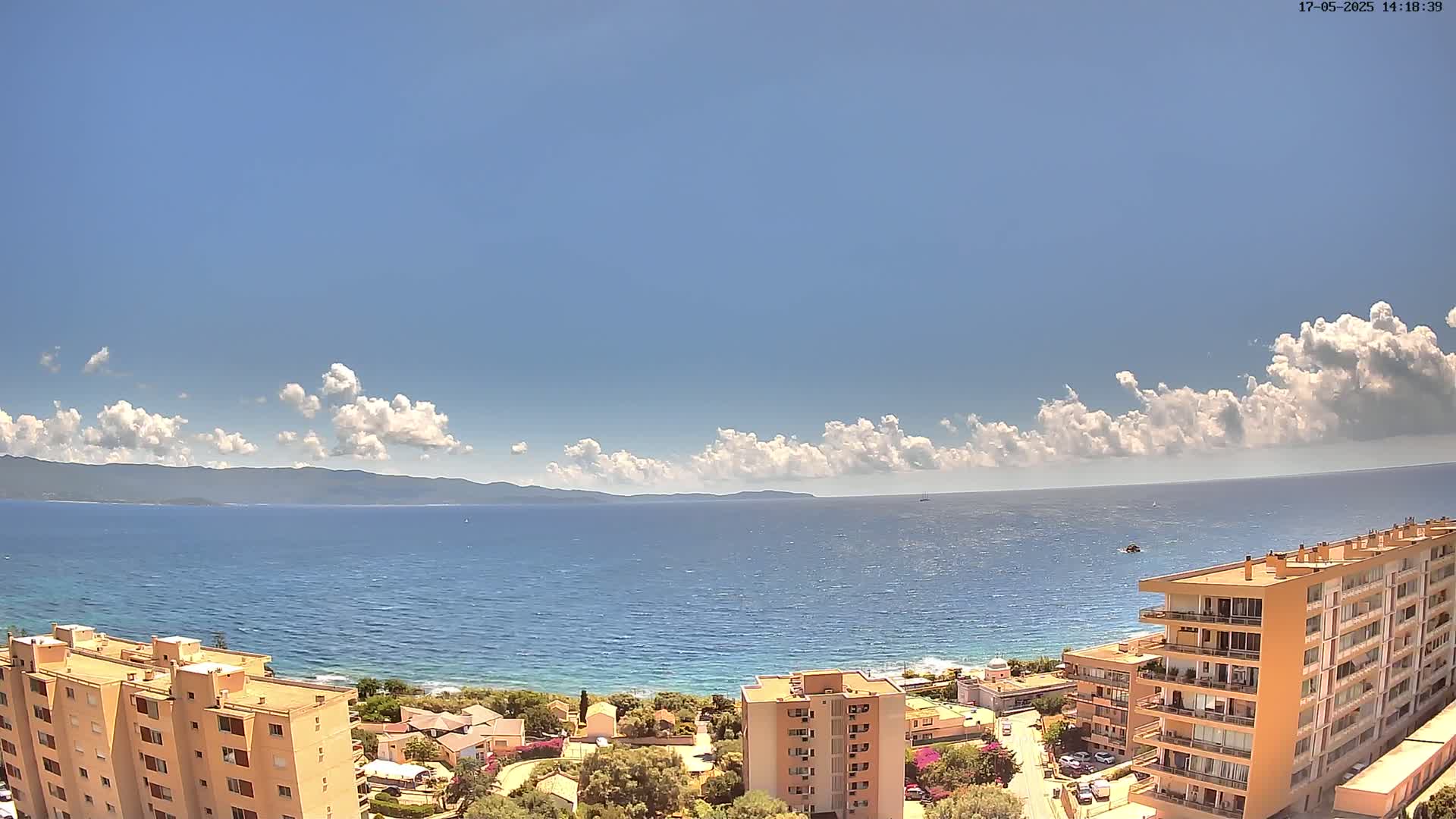 A partly cloudy, sunny day overlooks a calm sea with a distant mountain range, and several apartment buildings are visible in the foreground.