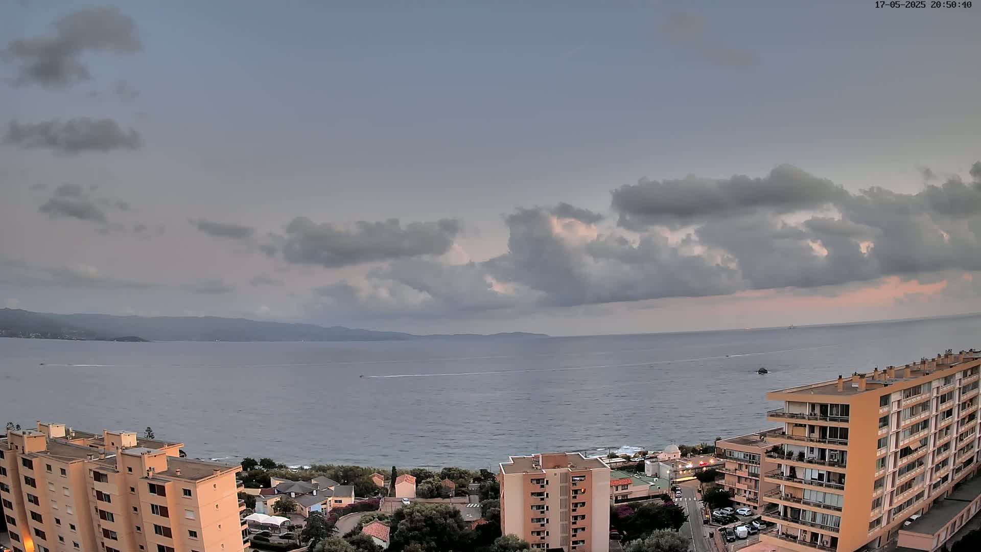 An elevated view shows a calm sea under a partly cloudy sky, with several apartment buildings situated along a coastline and hills visible in the distance.