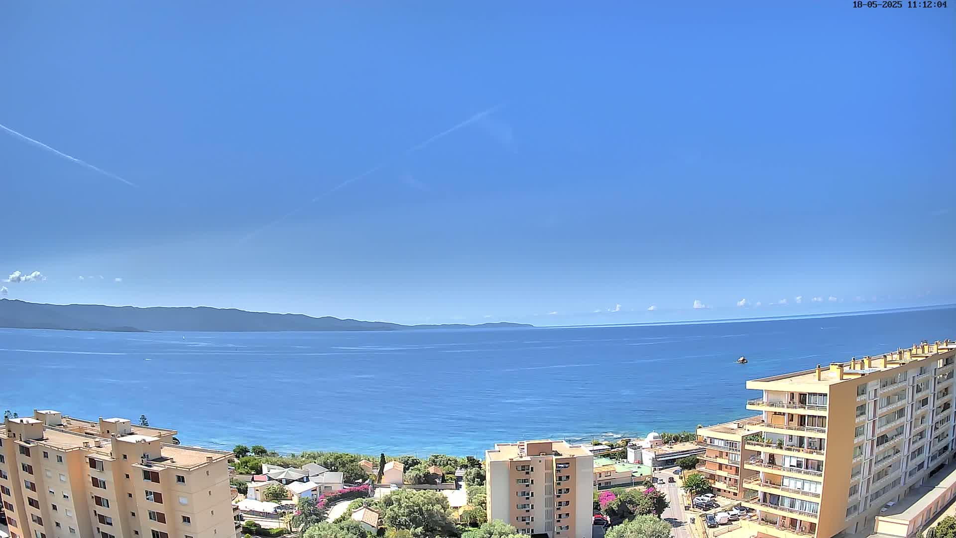 A clear blue sky overlooks a calm blue sea, a distant mountain range, and a coastal town with apartment buildings.