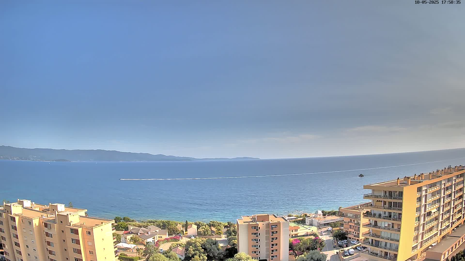 An aerial view shows a calm, blue sea under a mostly clear sky, with distant mountains and several apartment buildings in the foreground.