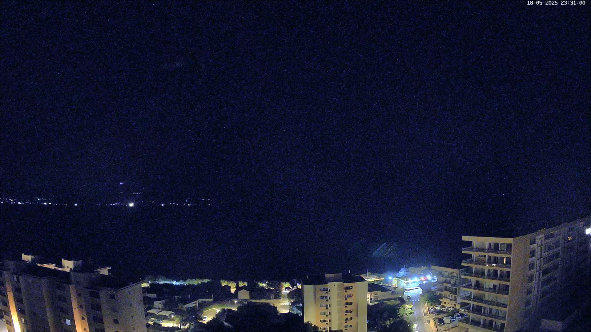 A nighttime, high-angle view shows several multi-story buildings illuminated against a dark, clear sky overlooking a body of water with faint lights on the opposite shore.