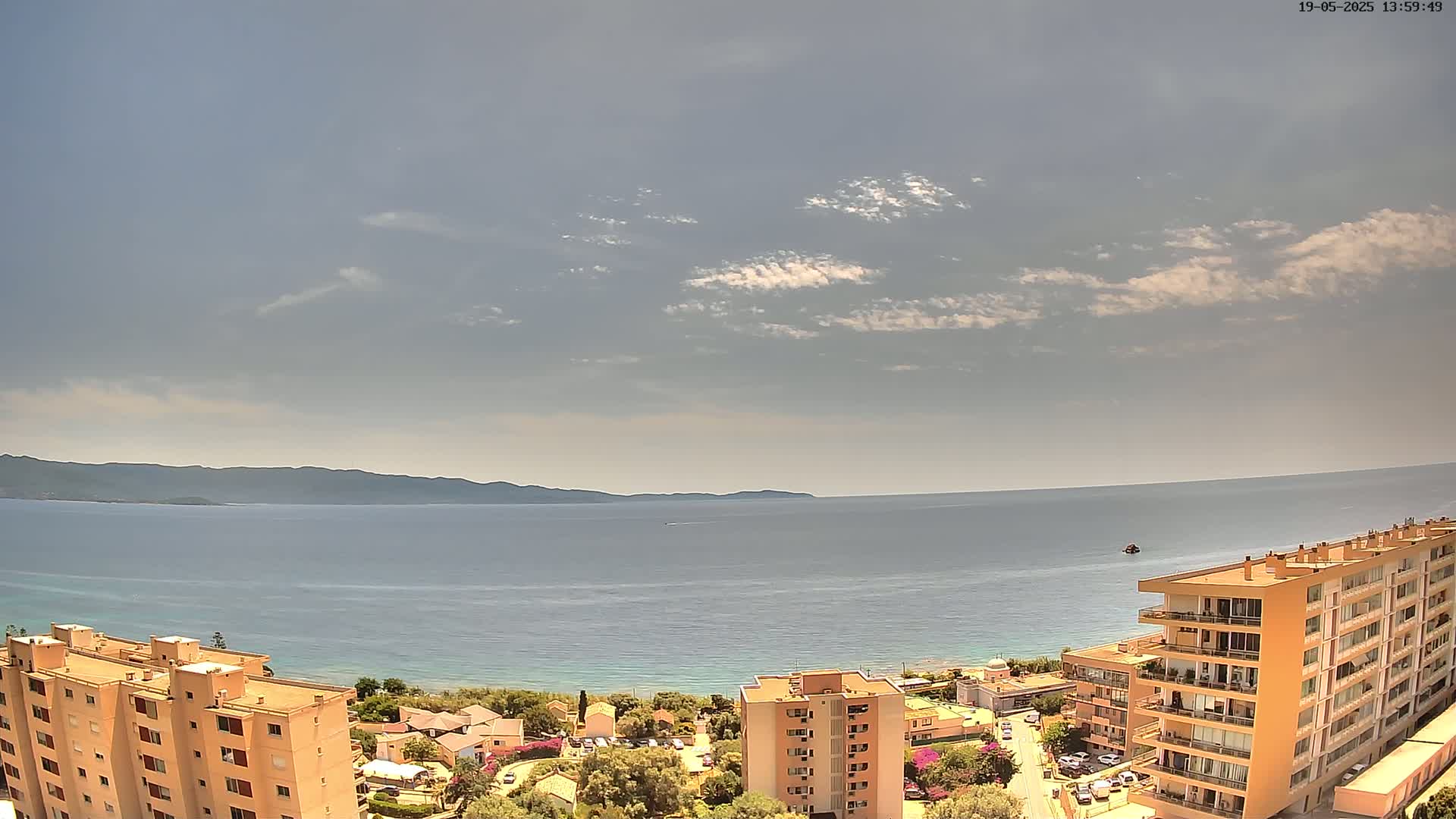 An aerial view shows a calm sea under a partly cloudy sky, with apartment buildings and coastal vegetation in the foreground.