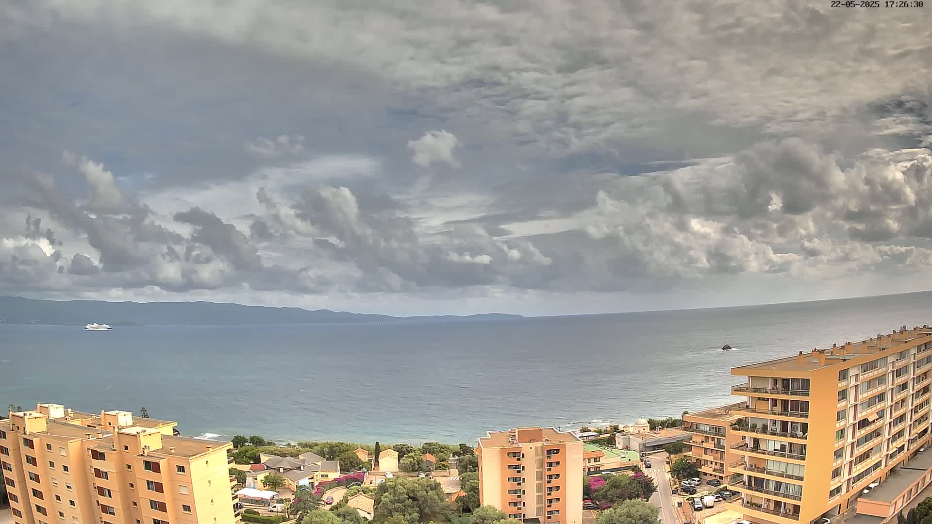 A partly cloudy day overlooks a calm sea with a cruise ship in the distance and several buildings along the coast.