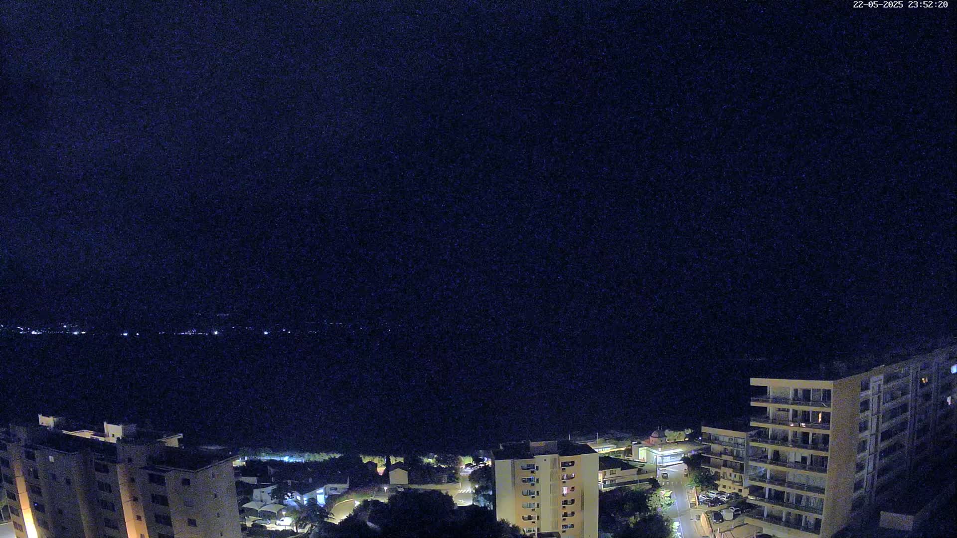 A nighttime view of a coastal city with apartment buildings, a dark body of water, and a clear sky.
