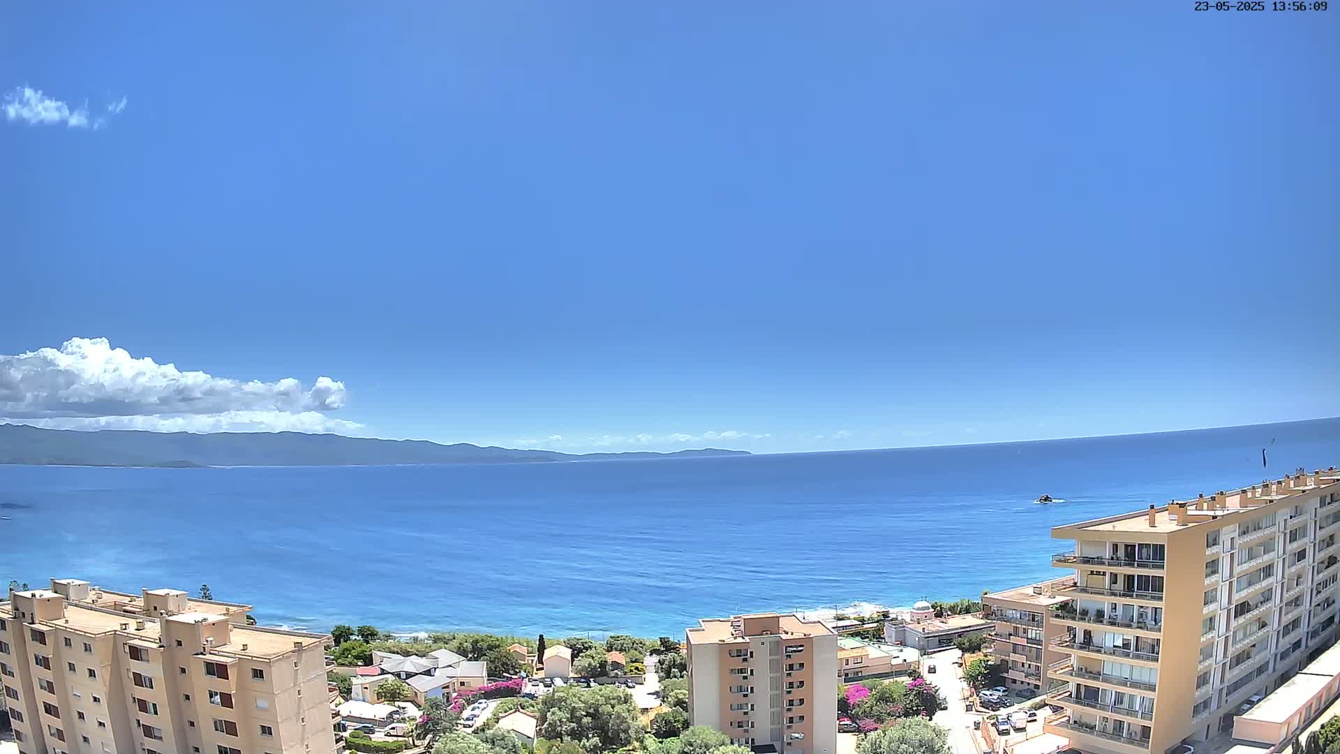 A mostly sunny day overlooking a calm blue ocean, with a distant mountain range and several multi-story buildings in the foreground.