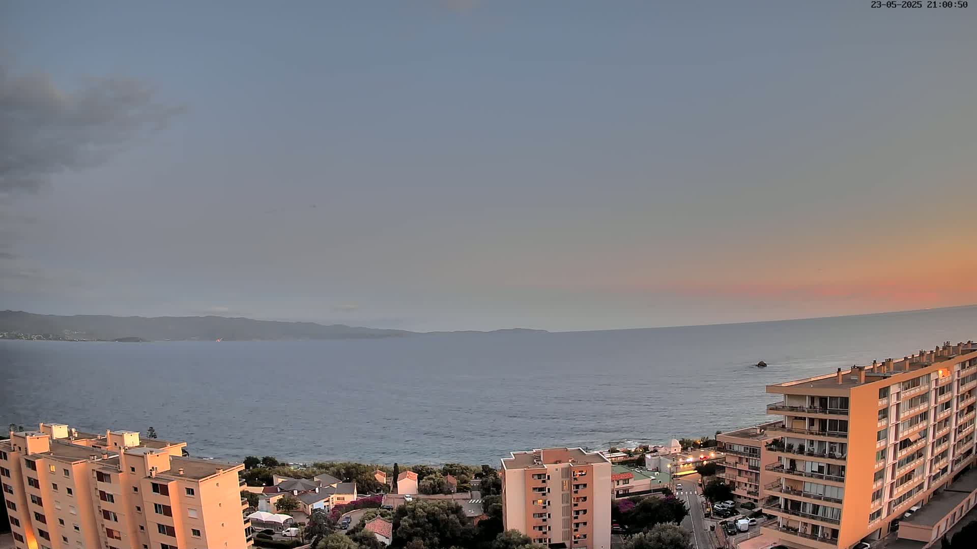 A calm sea at sunset is viewed from above, showing a coastline with apartment buildings and a mountainous background under a mostly clear sky.