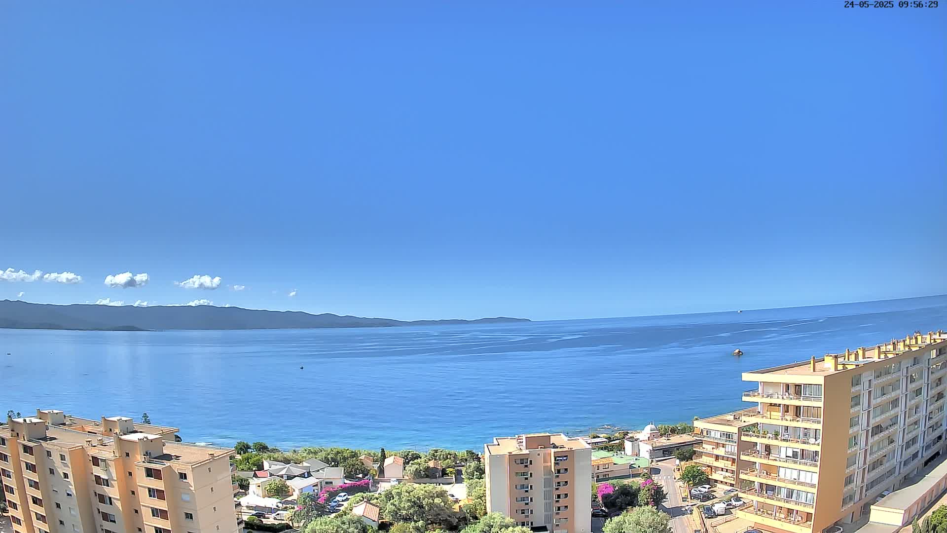 A mostly clear blue sky overlooks a calm blue sea, with a distant mountain range and several apartment buildings along the coast.