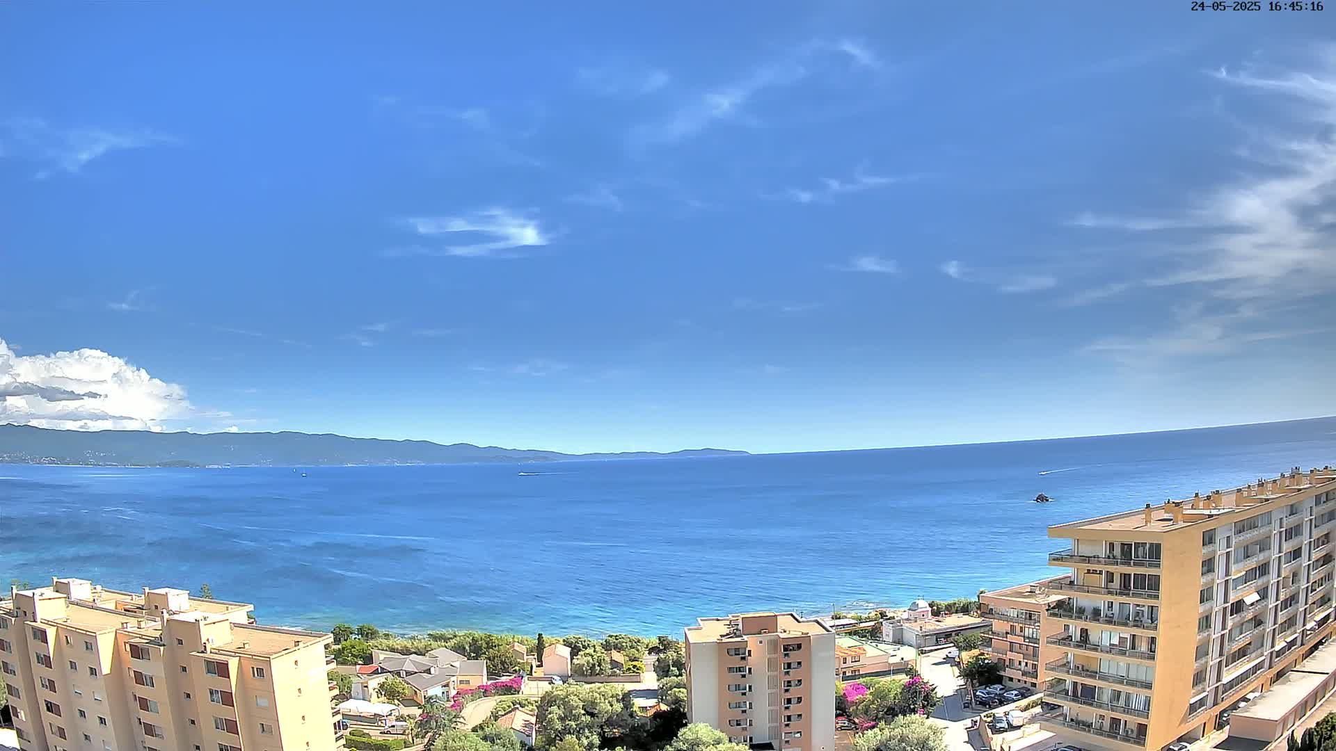 A mostly sunny day reveals a panoramic view of a calm blue sea, bordered by a distant mountain range and buildings along the coastline.