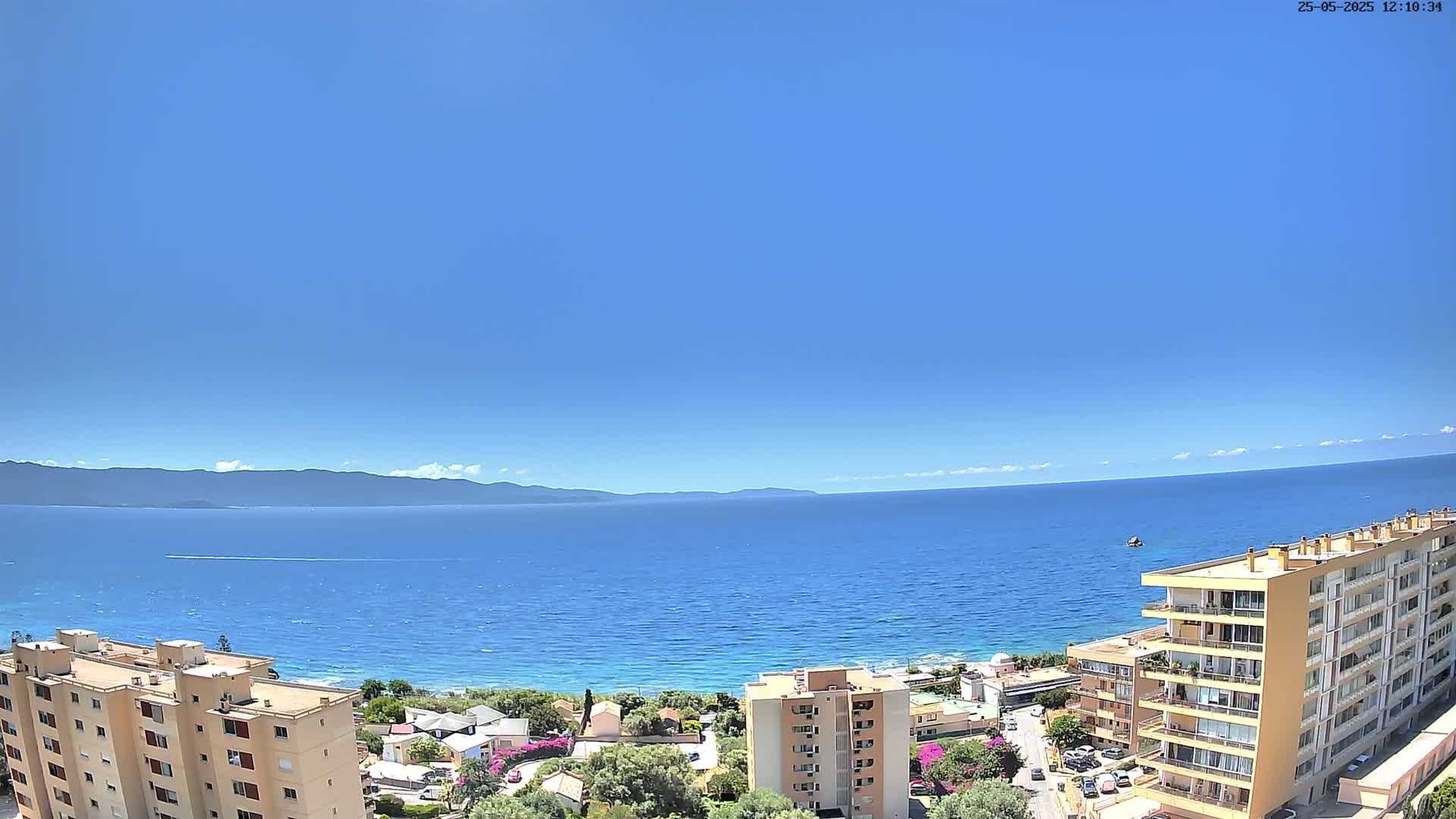 A clear blue sky overlooks a calm blue sea, with a distant mountain range visible on the horizon and apartment buildings in the foreground.