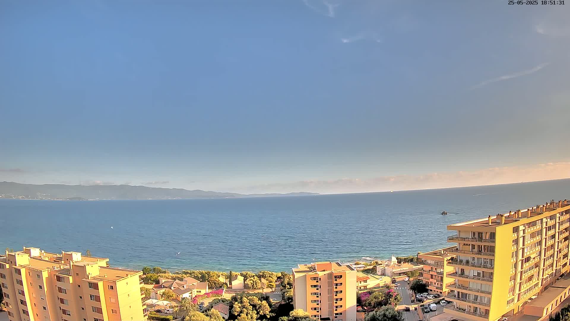 A coastal view shows a calm sea under a clear blue sky, with several multi-story buildings situated on a hillside overlooking the water and distant mountains.