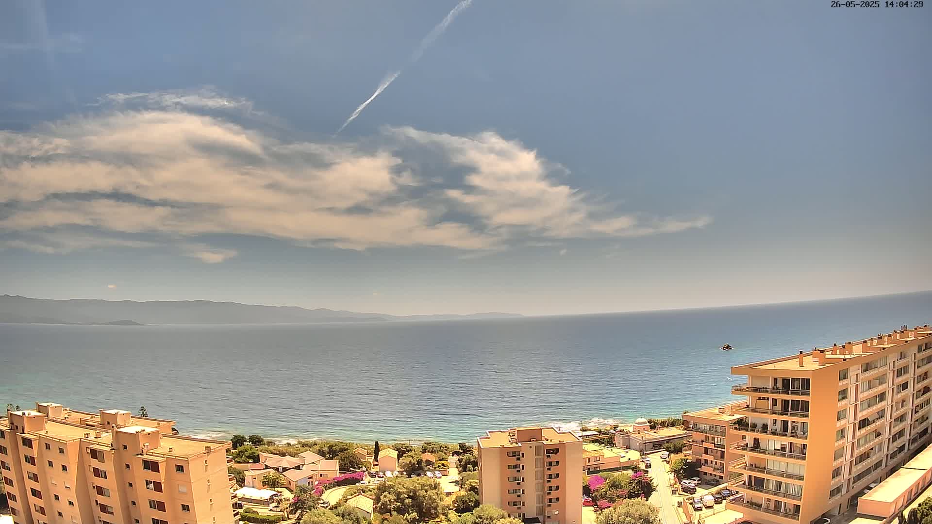 A mostly sunny day overlooks a calm ocean and coastline dotted with buildings, with a distant mountain range visible in the background.