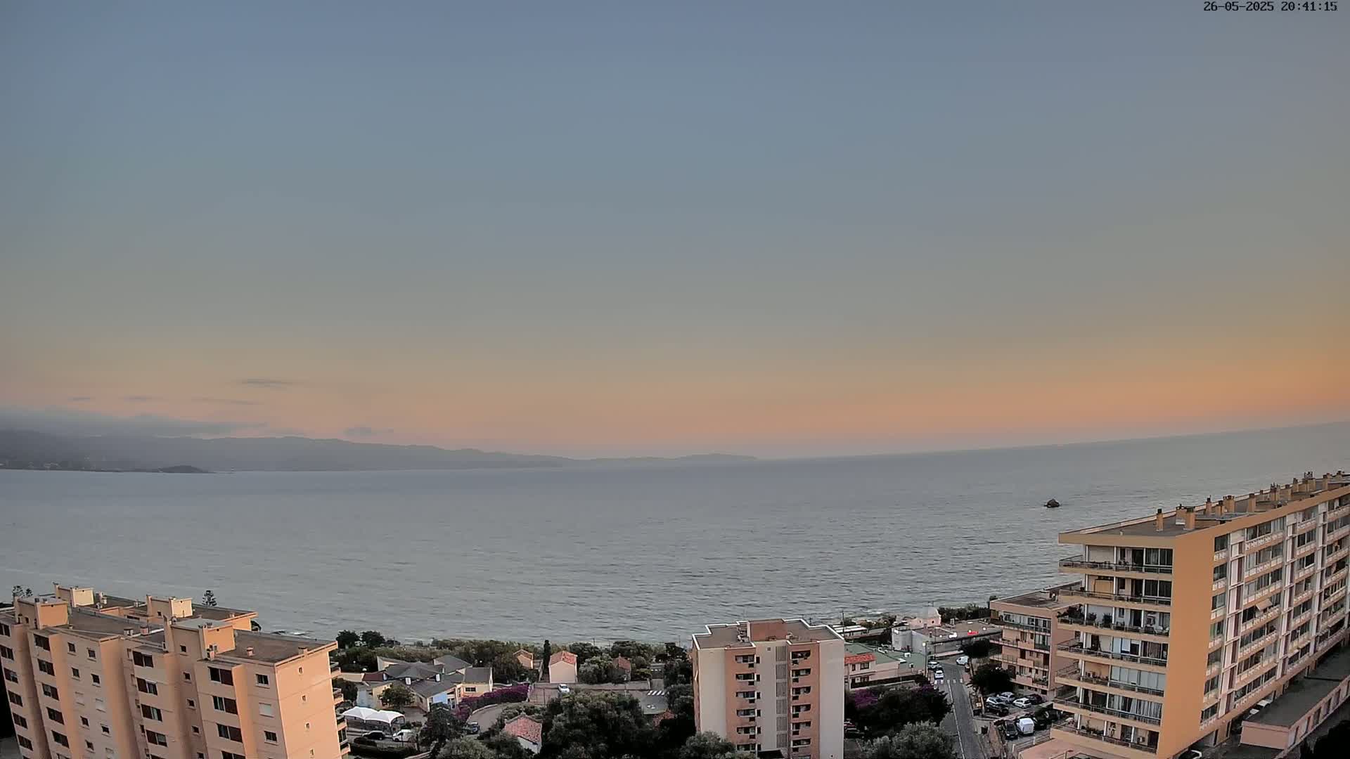 An elevated view of a calm sea at sunset, with apartment buildings situated along the coast and mountains visible in the distance.