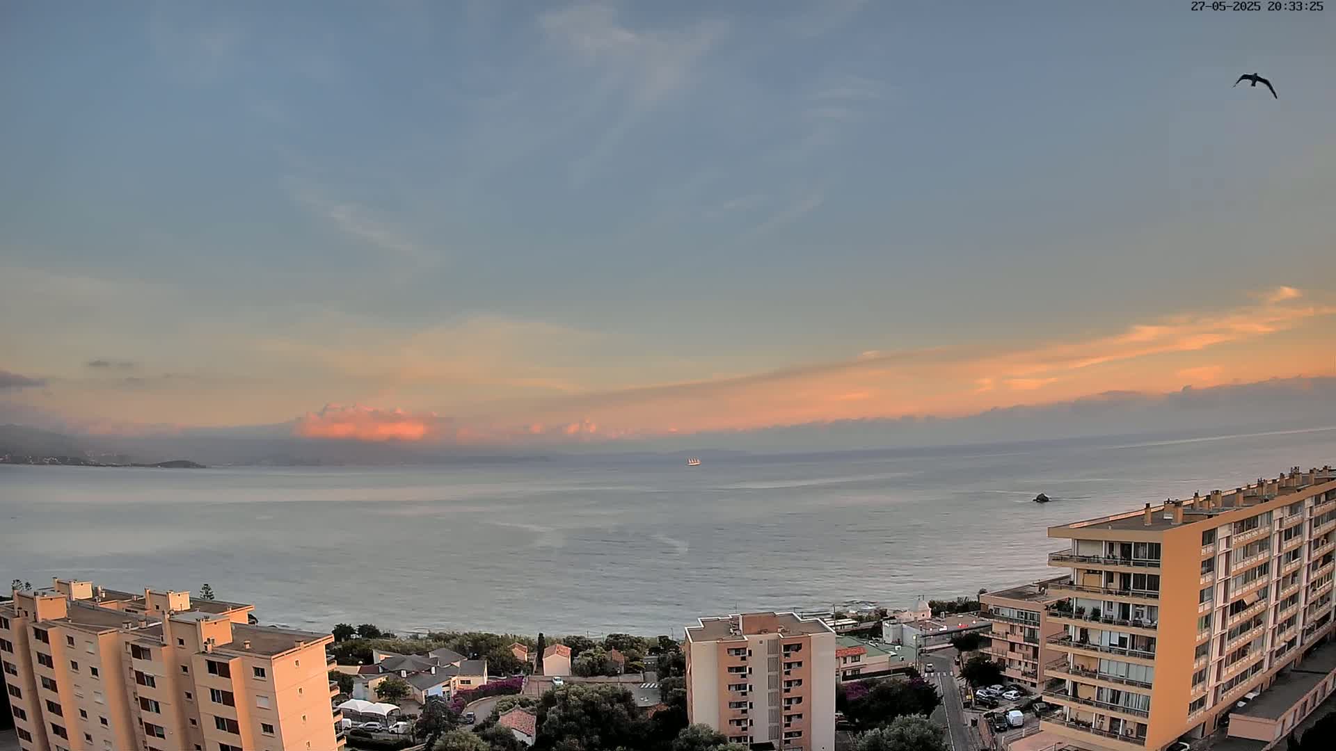 A calm ocean at sunset is viewed from above, showing a coastline with apartment buildings and a single bird in flight under a mostly clear sky.
