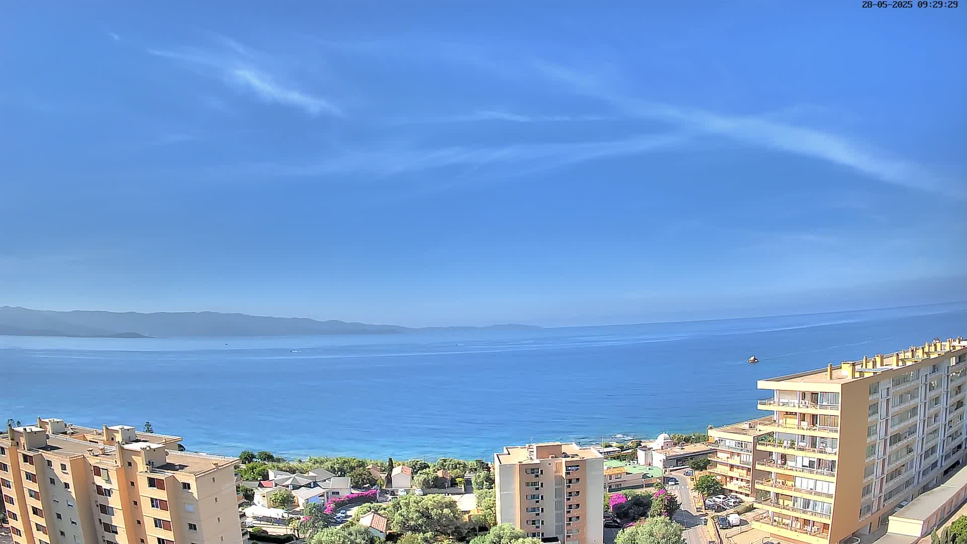 A mostly clear blue sky overlooks a calm blue sea, with distant mountains and several apartment buildings visible near the shore.