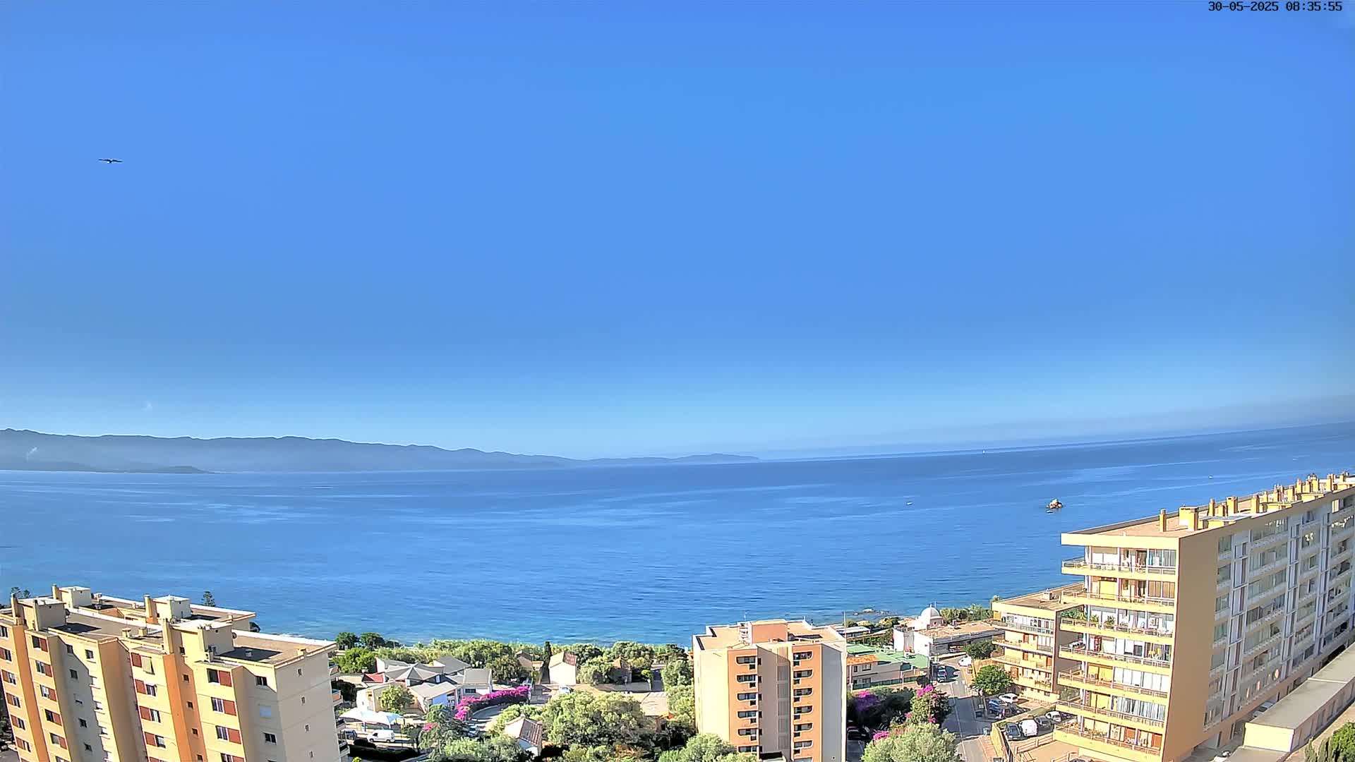 A clear blue sky overlooks a calm blue sea, mountains in the distance, and several multi-story buildings along a coastline with some vegetation.