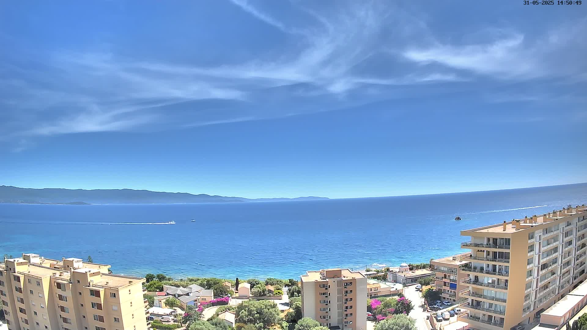 Under a mostly clear, bright blue sky, a coastal scene shows a calm blue sea extending to a distant mountain range, with several apartment buildings and residential areas visible in the foreground.