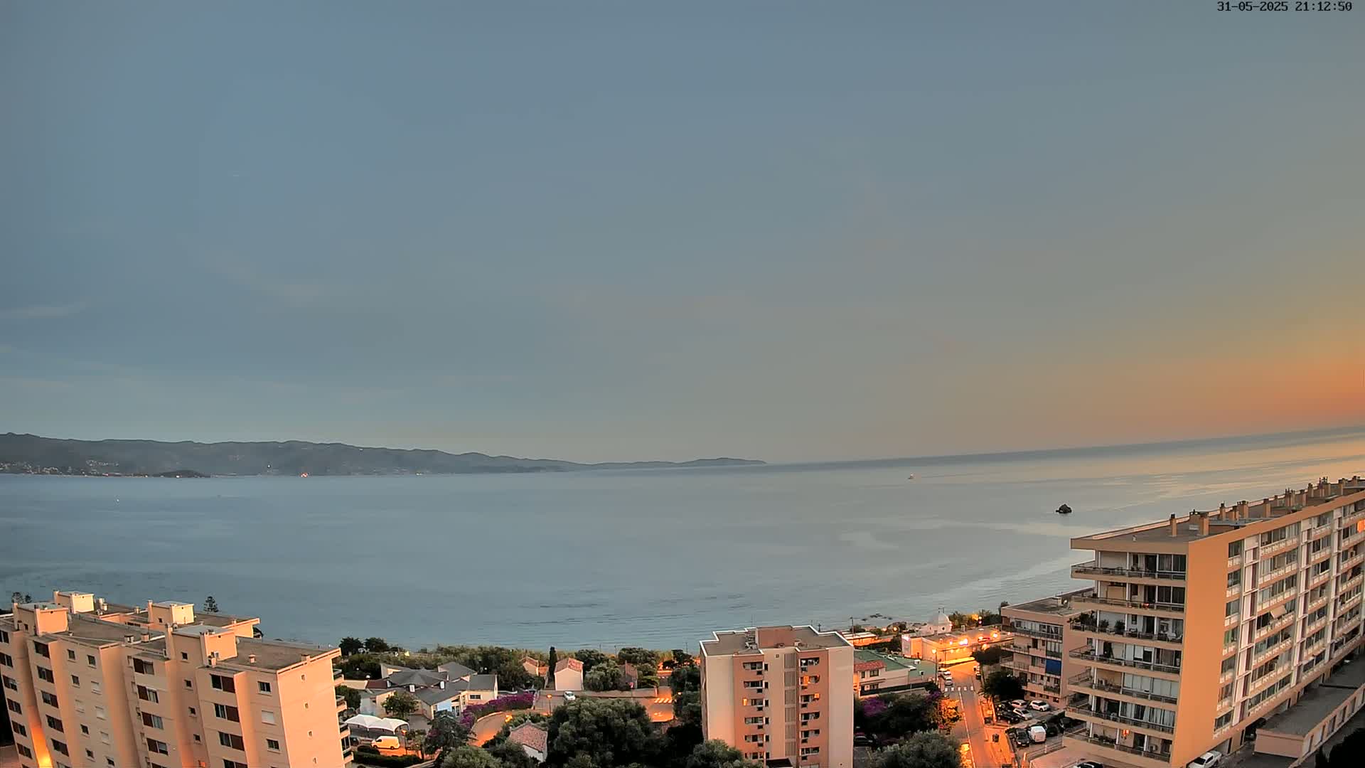 A calm sea under a pale sunset sky is viewed from a high vantage point overlooking a coastal town with several apartment buildings.