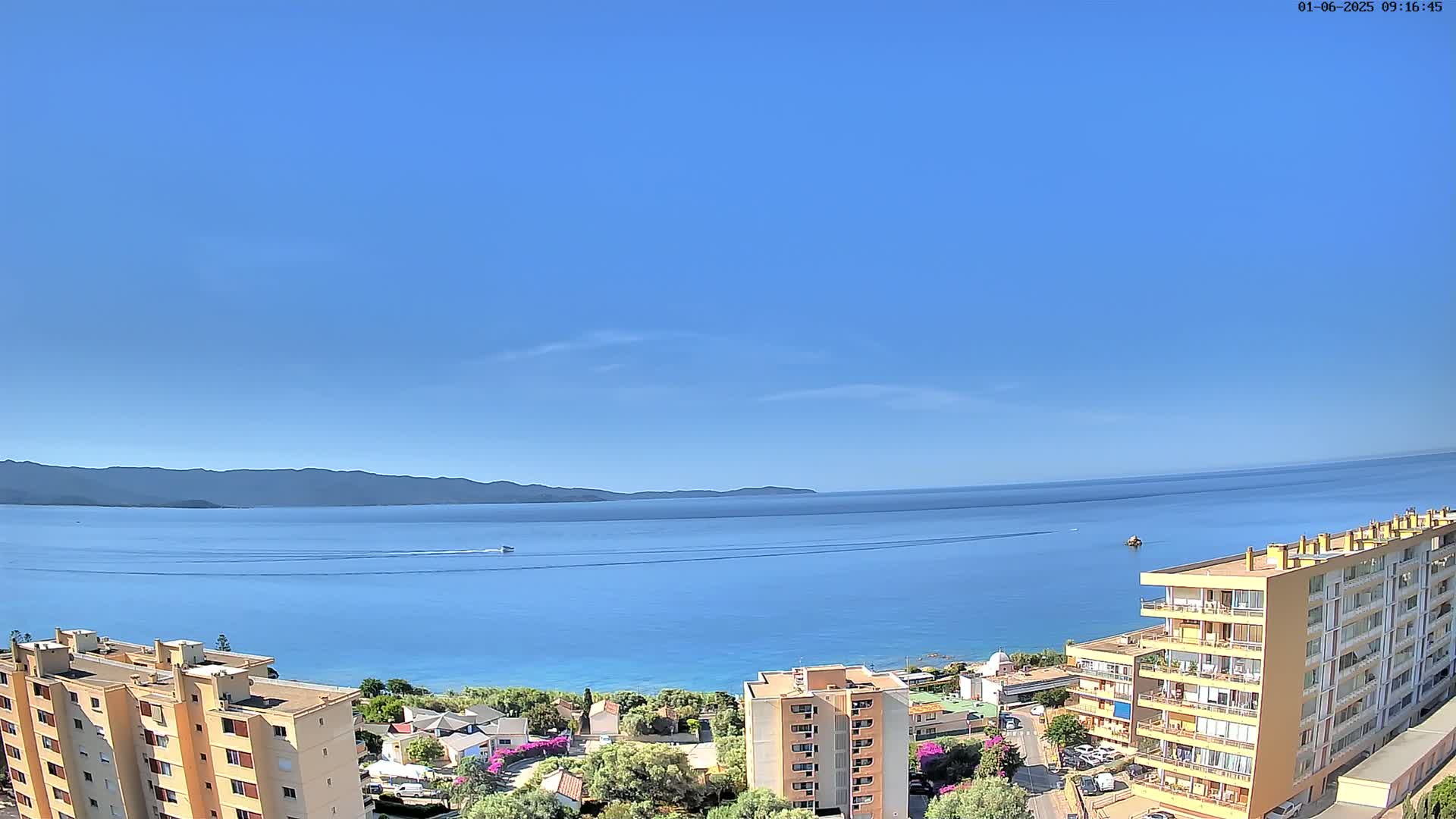 A clear blue sky overlooks a calm blue sea with distant mountains, and several apartment buildings are situated on the shore.