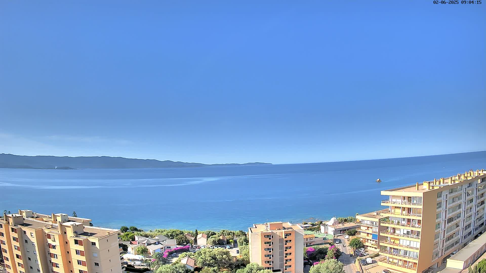 A clear, sunny day overlooking a calm blue sea and distant mountains, with several apartment buildings situated on a hillside overlooking the water.