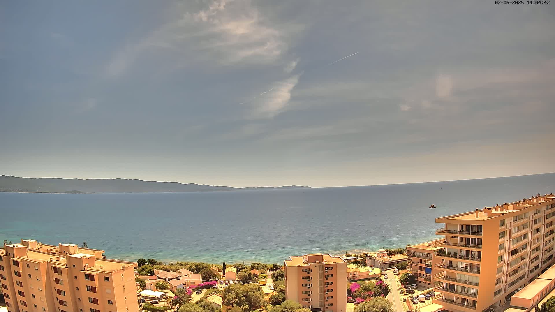 A partly cloudy sky overlooks a calm blue sea, with a coastal town and several apartment buildings in the foreground.