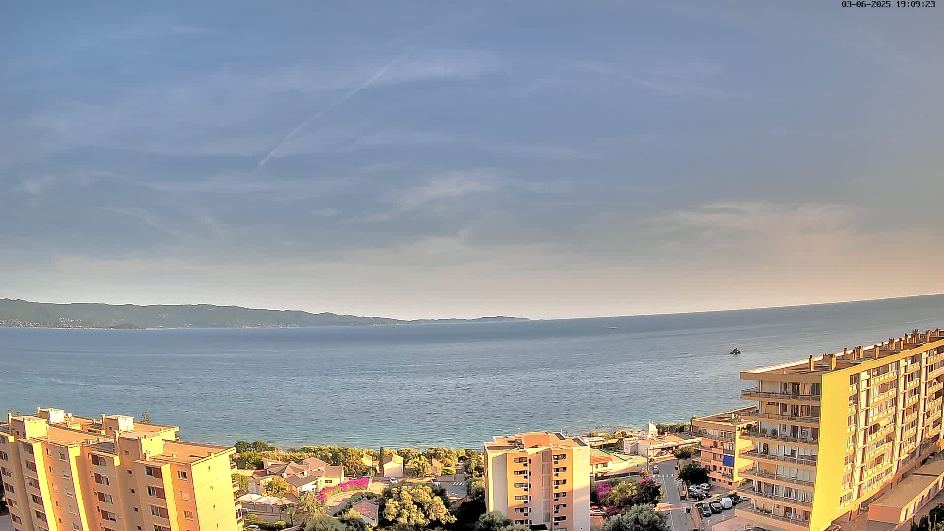 A mostly clear sky overlooks a calm sea with a distant shoreline, and several apartment buildings are situated closer to the viewer.