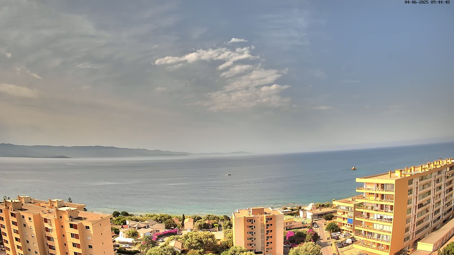 A partly cloudy day overlooks a calm sea with a distant mountain range and several apartment buildings near the shore.