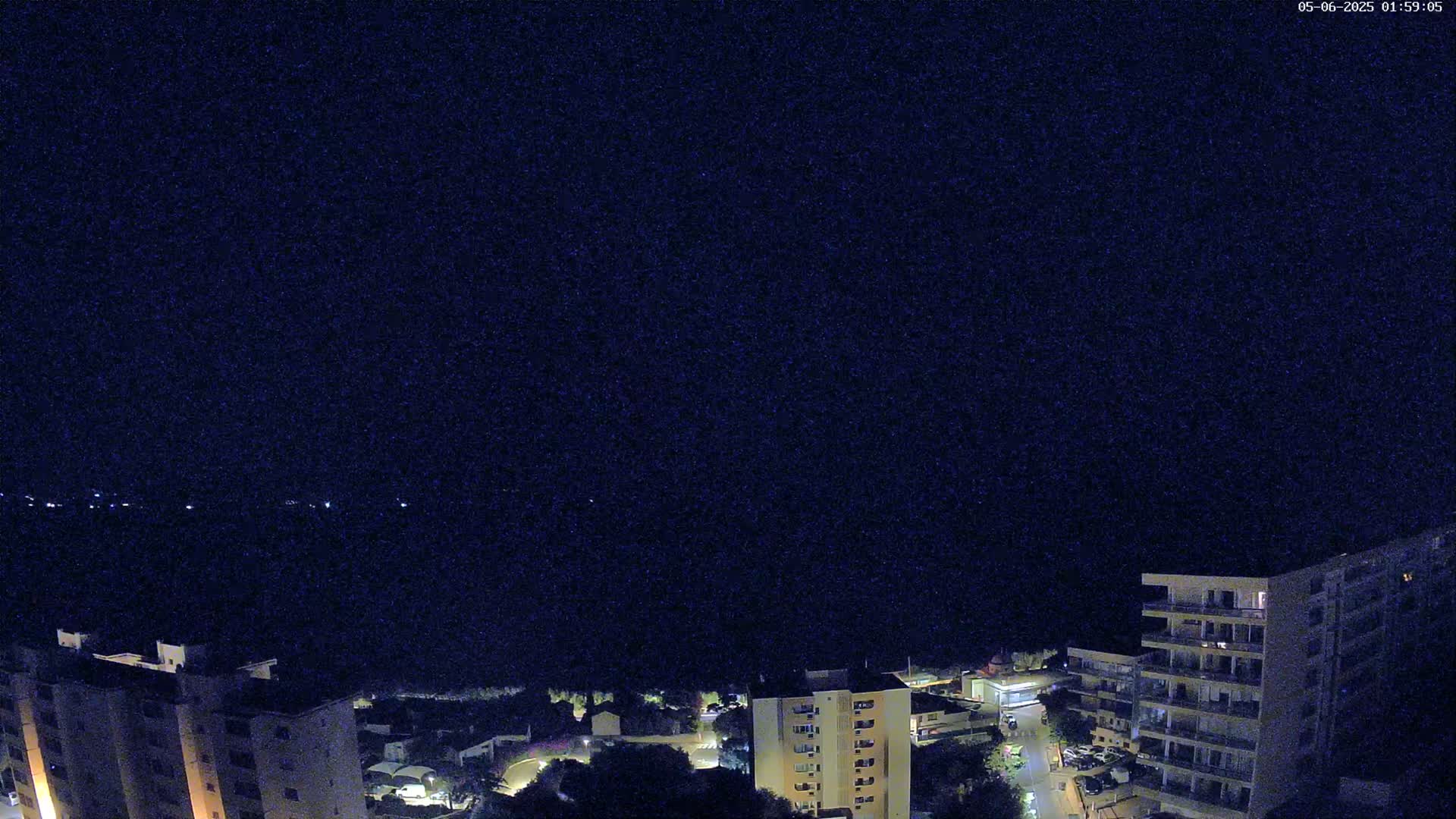 A nighttime, high-angle view shows a coastal town with several apartment buildings illuminated, overlooking a dark body of water under a clear, dark sky.