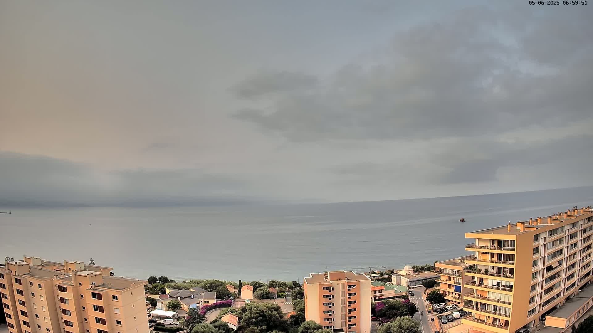 A coastal town with apartment buildings is visible under an overcast sky, overlooking a calm sea.