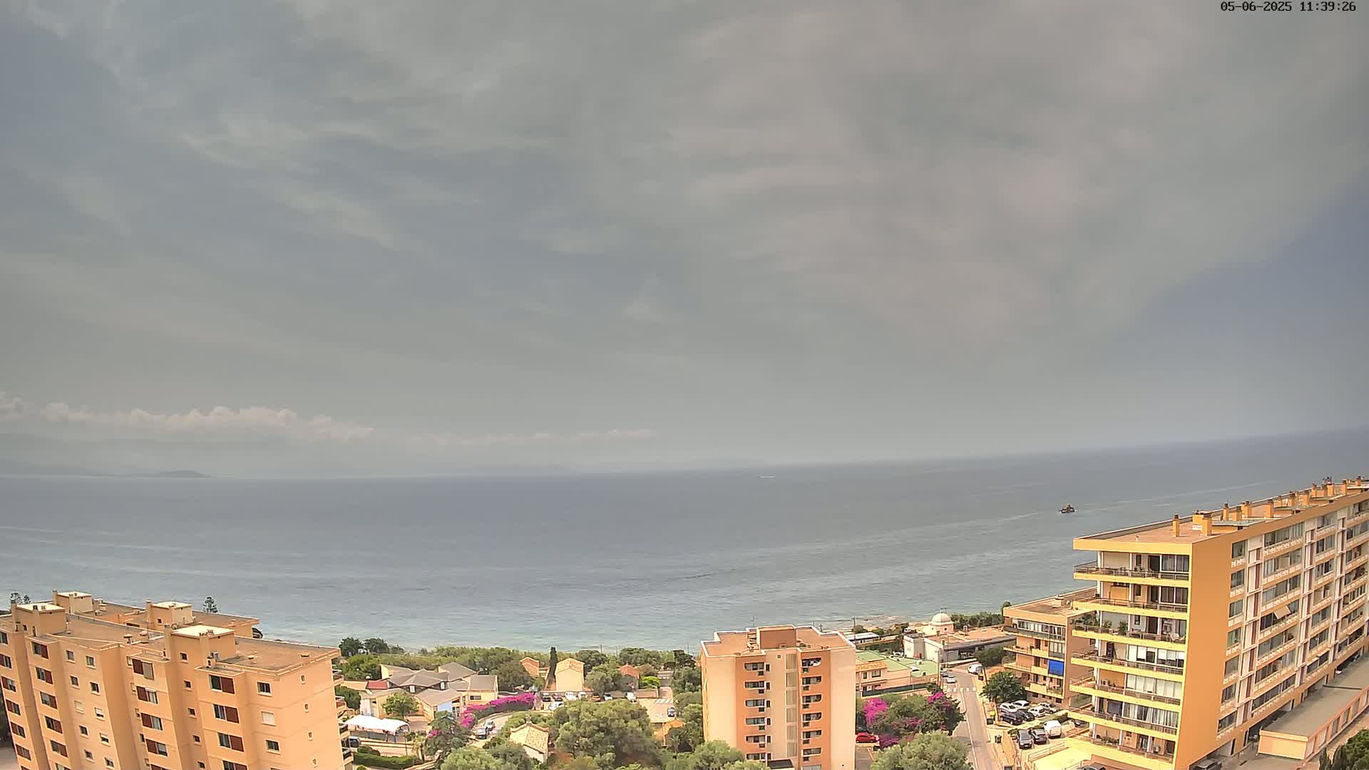 An overcast sky hangs over a calm sea, overlooking a coastal town with several apartment buildings and greenery.