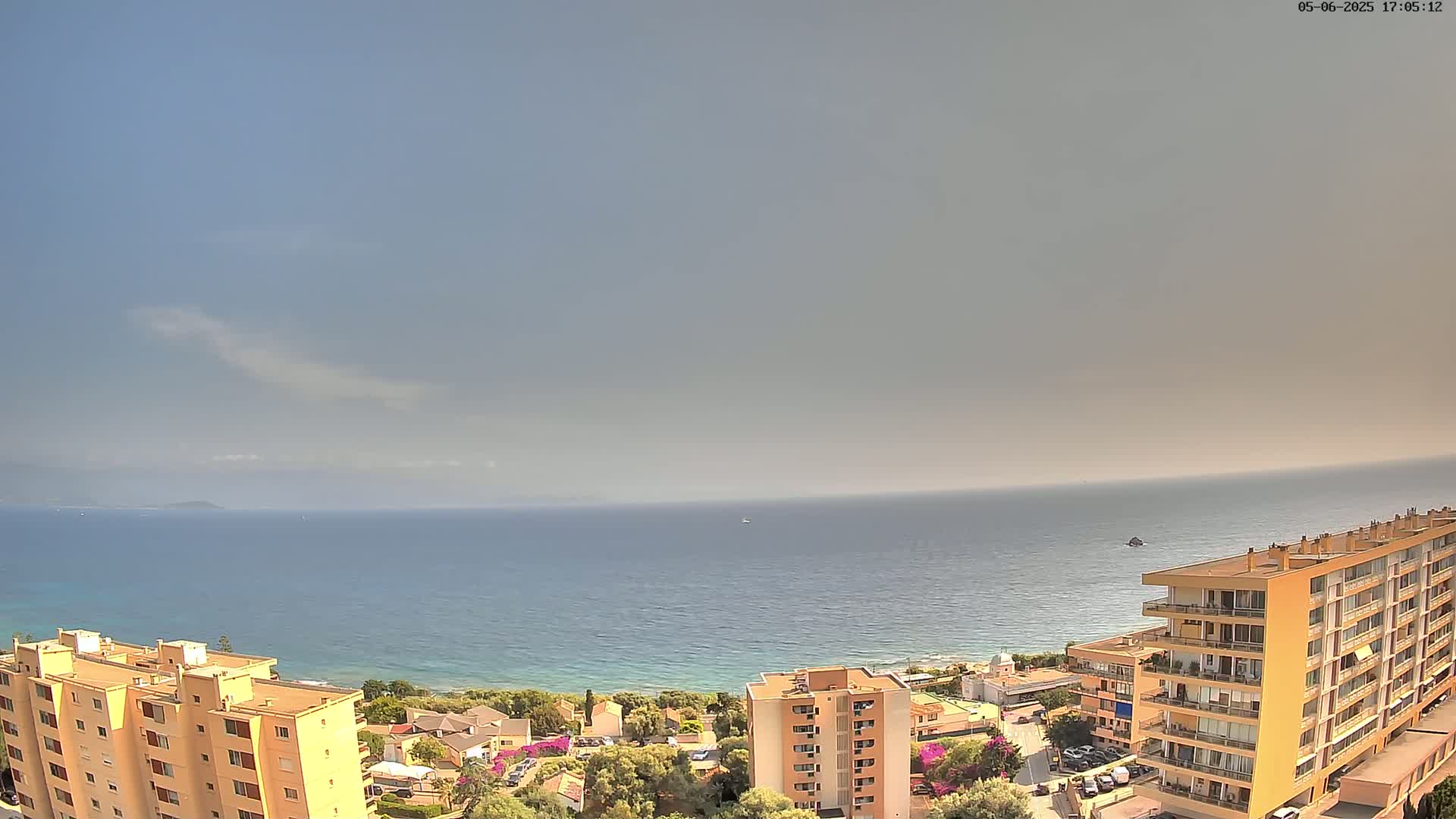 A hazy, sunny day overlooks a calm blue sea, with several apartment buildings and green vegetation situated on a coastal hillside.